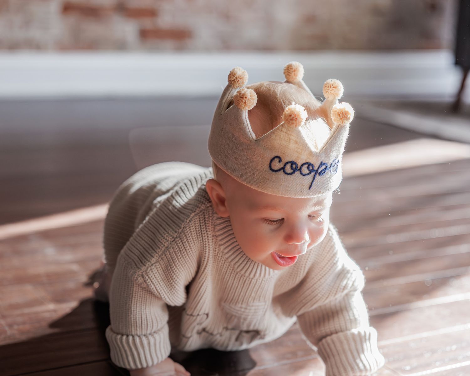 Baby boy named Cooper crawls on hardwood floor wearing beige knit outfit and personalized crown hat.