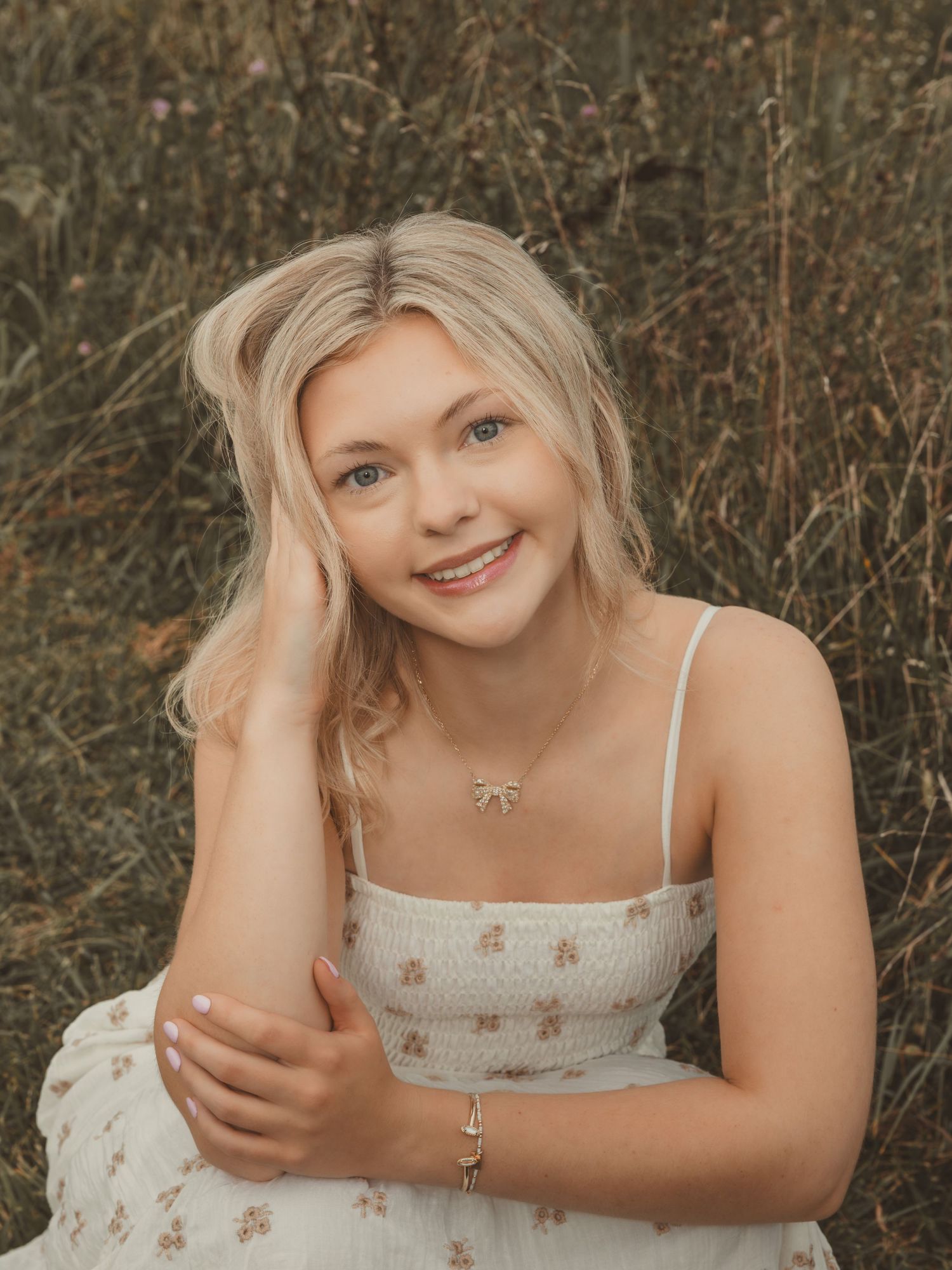 Smiling blonde teenage girl in white floral dress sitting outdoors in a field for senior portrait photo.