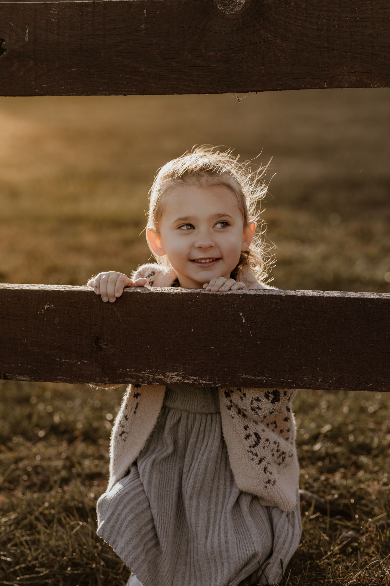 A small child peeks playfully over a dark wooden fence in warm sunlight.