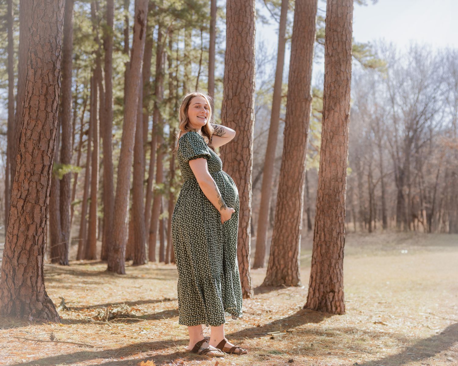 A maternity photoshoot in a sunlit forest with tall pine trees and a flowing green floral dress.