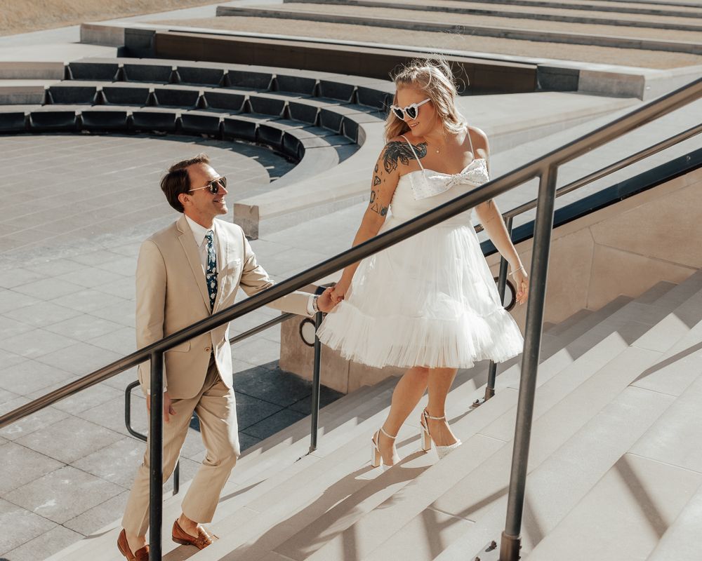 Bride in short white tulle dress and groom in tan suit walk hand-in-hand down sunny outdoor amphitheater steps.