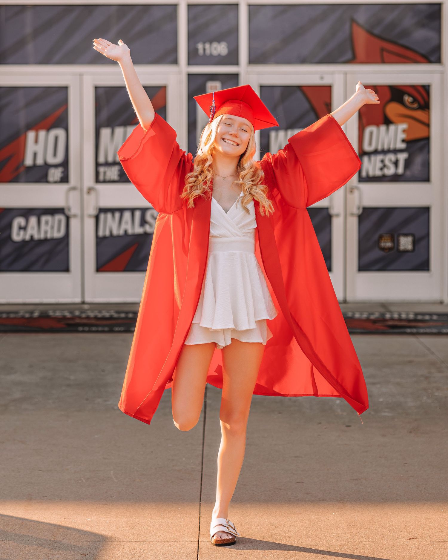 Joyful graduate in red cap and gown poses with arms raised outside Cardinals school building.