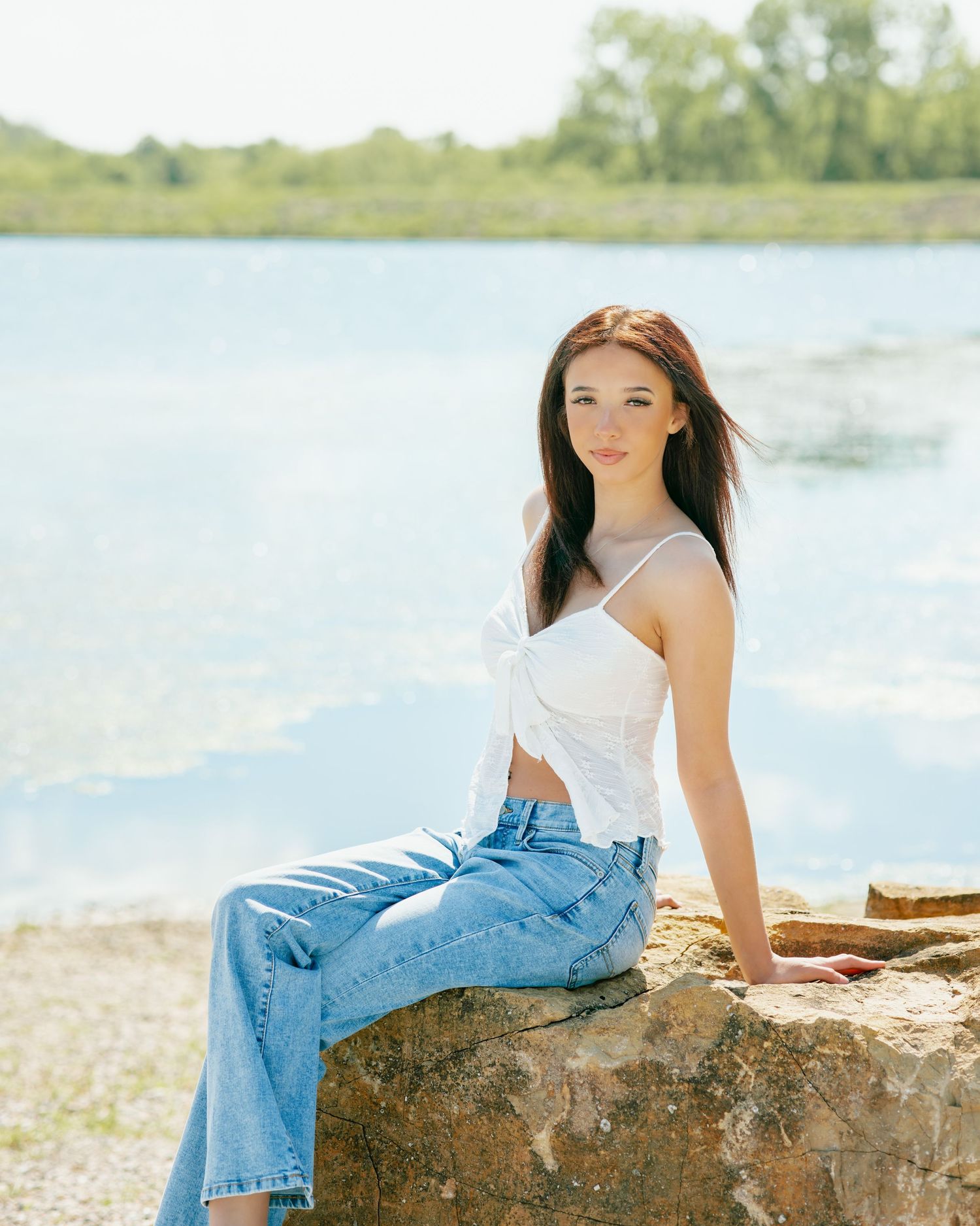 Young woman in white top and jeans sitting on rocks by a serene lake on a sunny day.