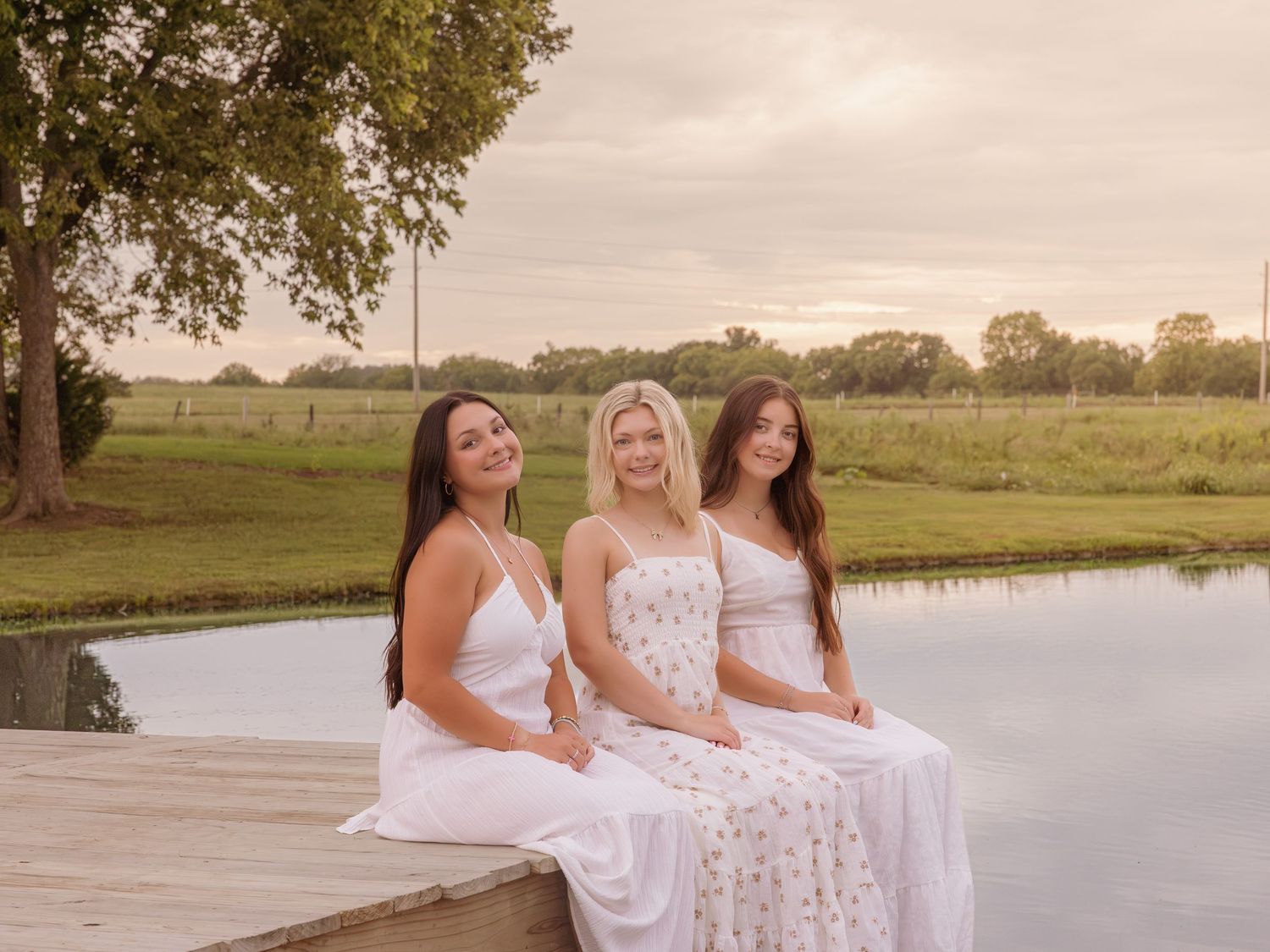 Three young women in white dresses sitting on a wooden dock by a serene pond surrounded by lush greenery.