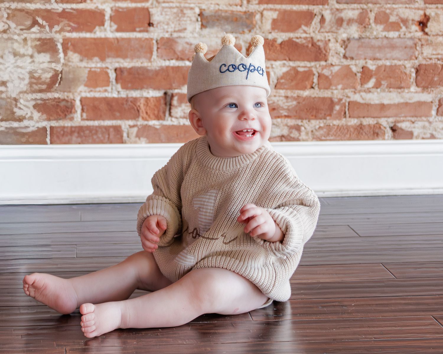 Smiling baby wearing a personalized crown hat sitting on hardwood floor against a brick wall.