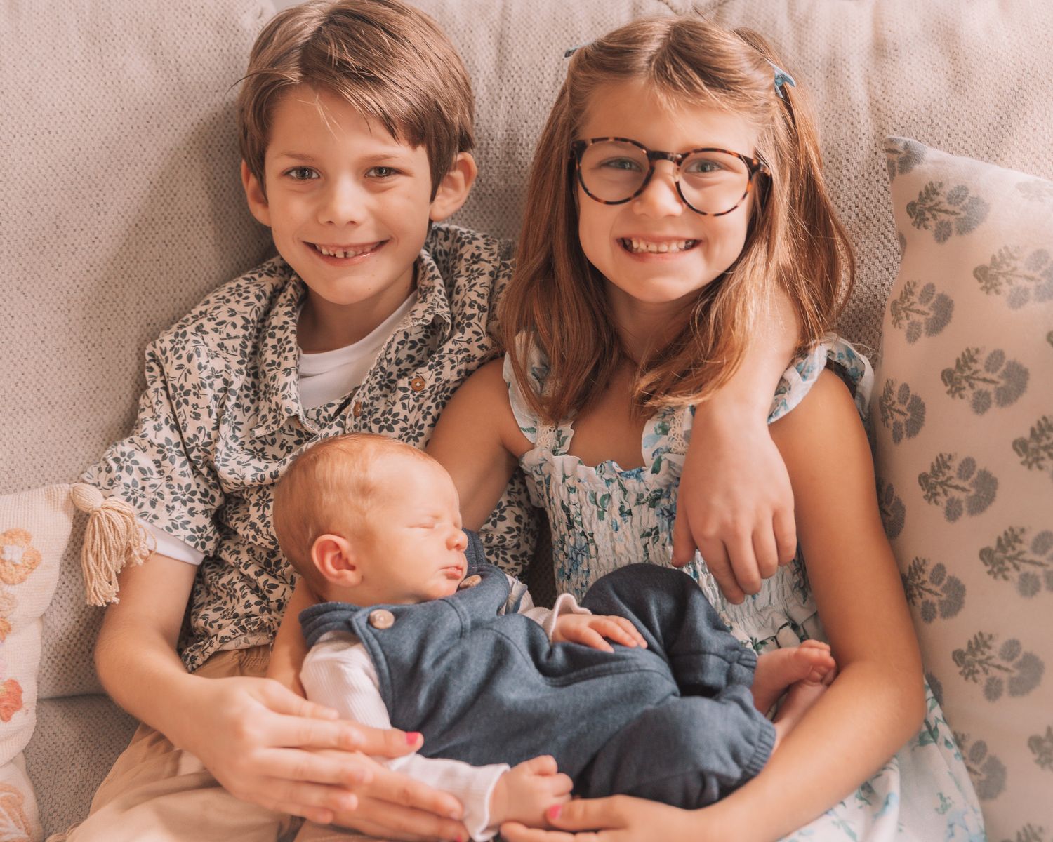Two smiling siblings, a boy and a girl with glasses, holding their newborn baby brother on a couch.