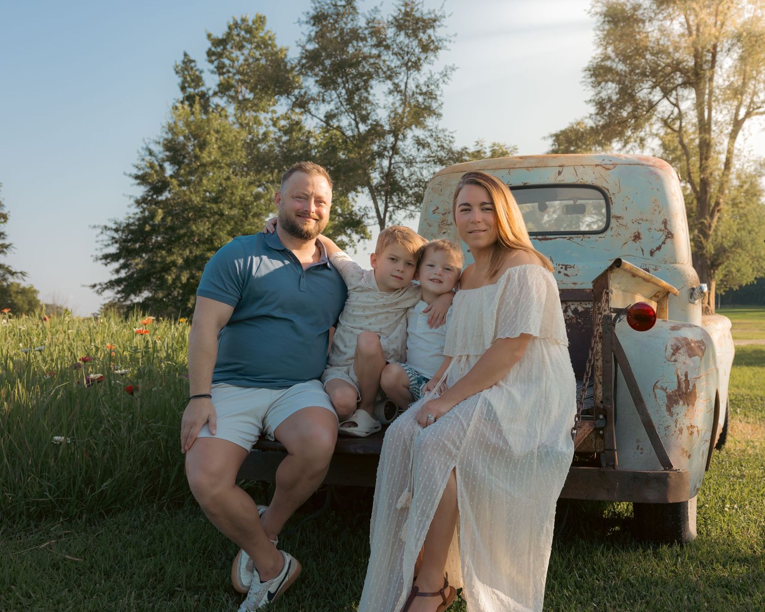 Family of four posing by a vintage rustic truck in a sunny green field during golden hour.