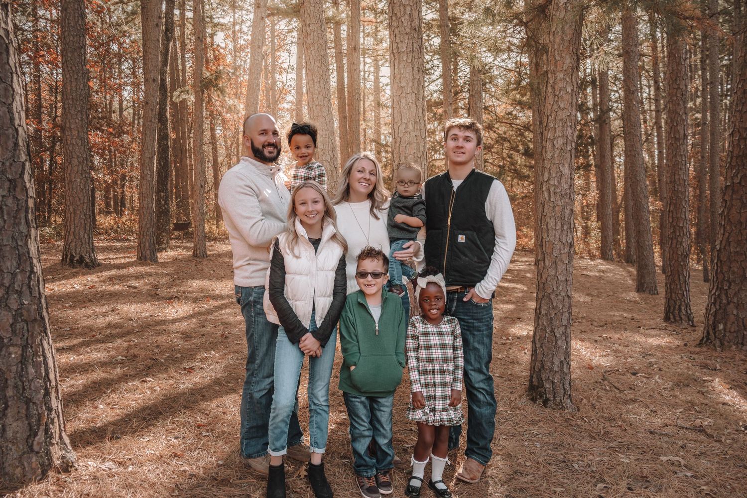 A group of people pose together in a sunlit forest with tall pine trees and brown needles covering the ground.