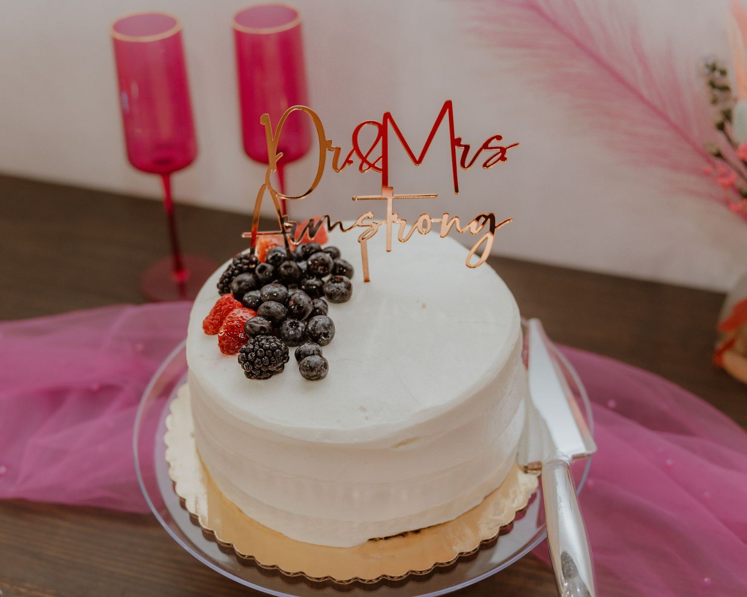 White wedding cake with Mr & Mrs Armstrong topper and fresh berries beside pink champagne flutes.