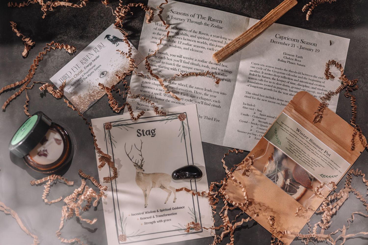 Mystical stag card, spell papers, crystals, and dried herbs arranged on a dark surface for a witchcraft subscription box.