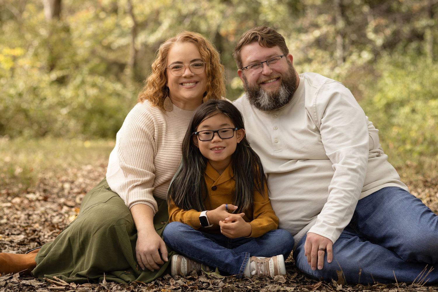 Family of three smiling outdoors in autumn, parents and young child with glasses sitting on fallen leaves.