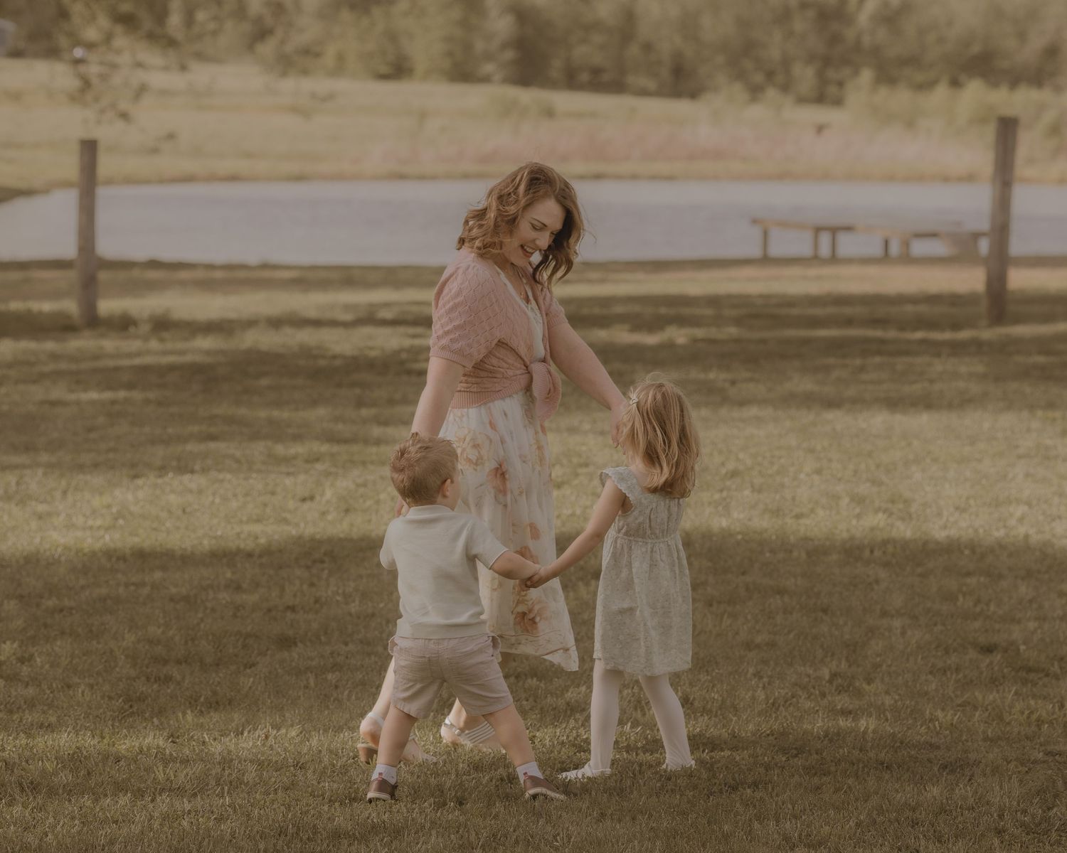 A joyful mother dances and holds hands with her two young children on a green lawn outdoors.
