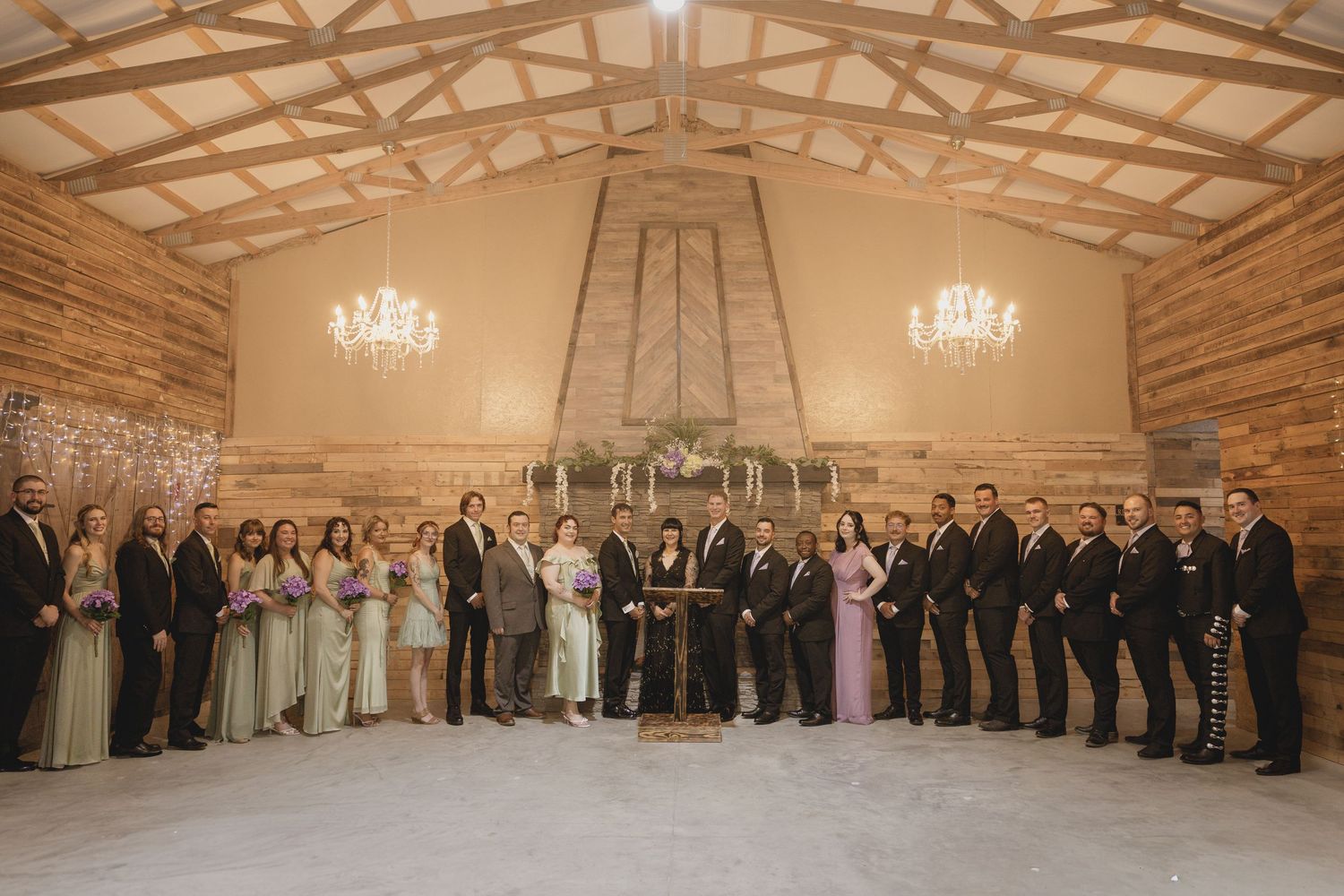 Large wedding party poses inside rustic barn venue with chandeliers, bridesmaids in sage green and groomsmen in black suits.