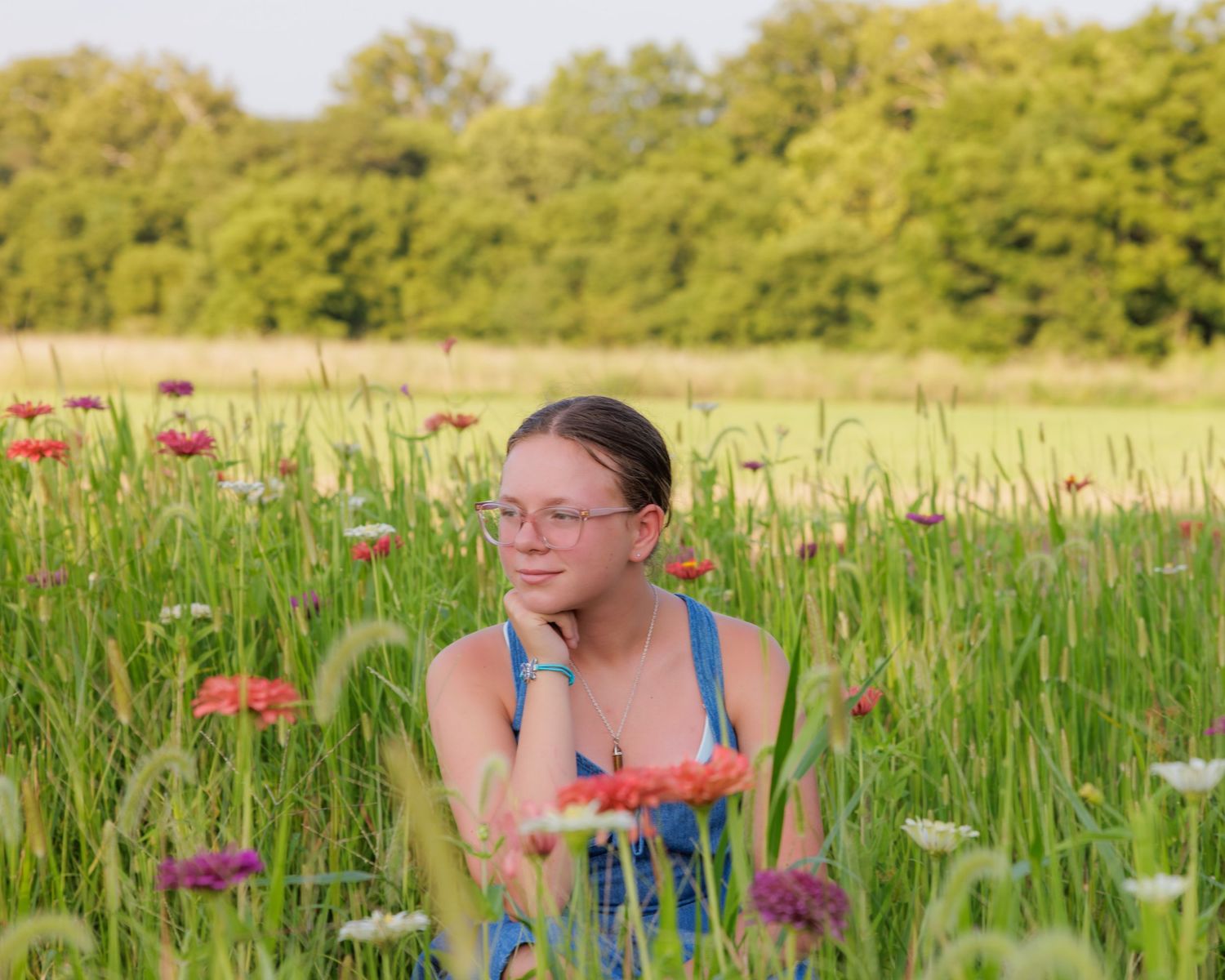 Young girl with glasses sitting in a colorful wildflower meadow, holding red flowers, with green trees in the background.