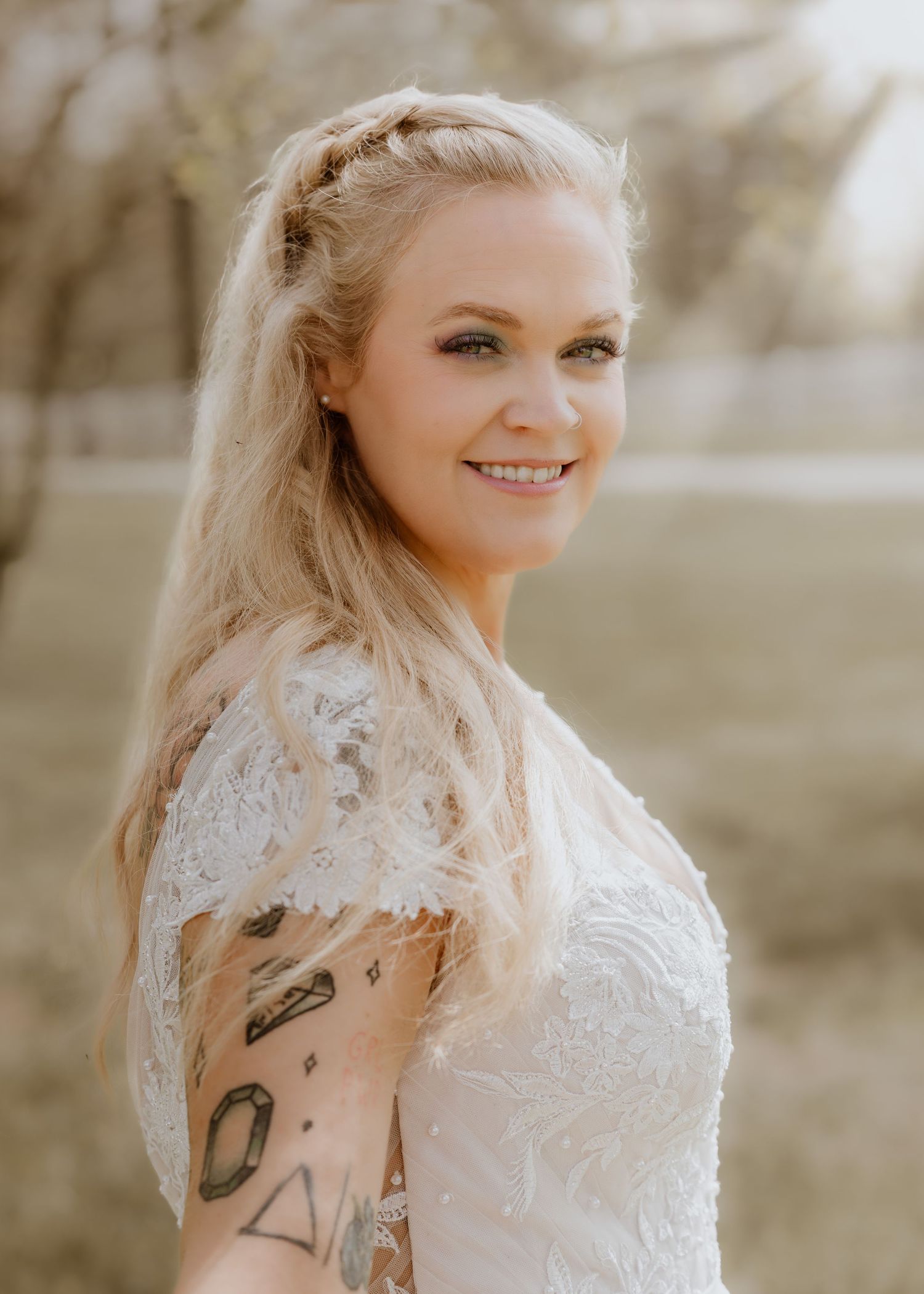 Blonde bride in lace wedding dress with braided updo and arm tattoos smiling outdoors.