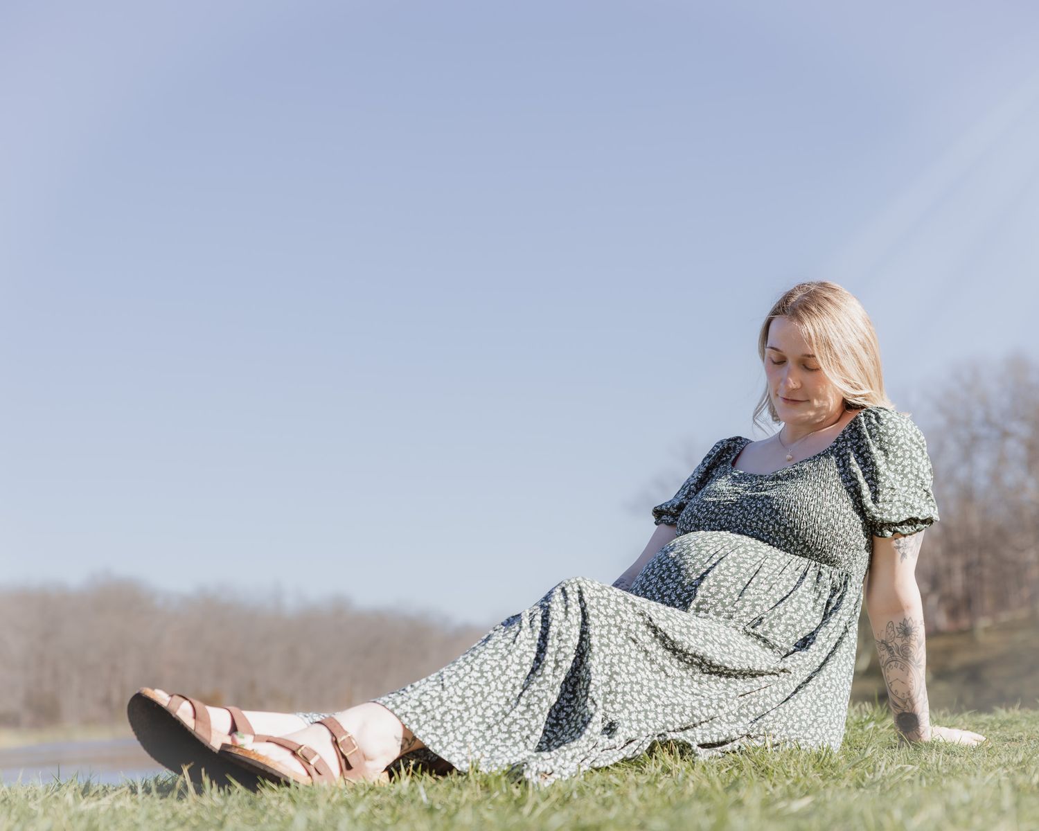 Pregnant woman in floral dress relaxing on grass outdoors on a sunny day.