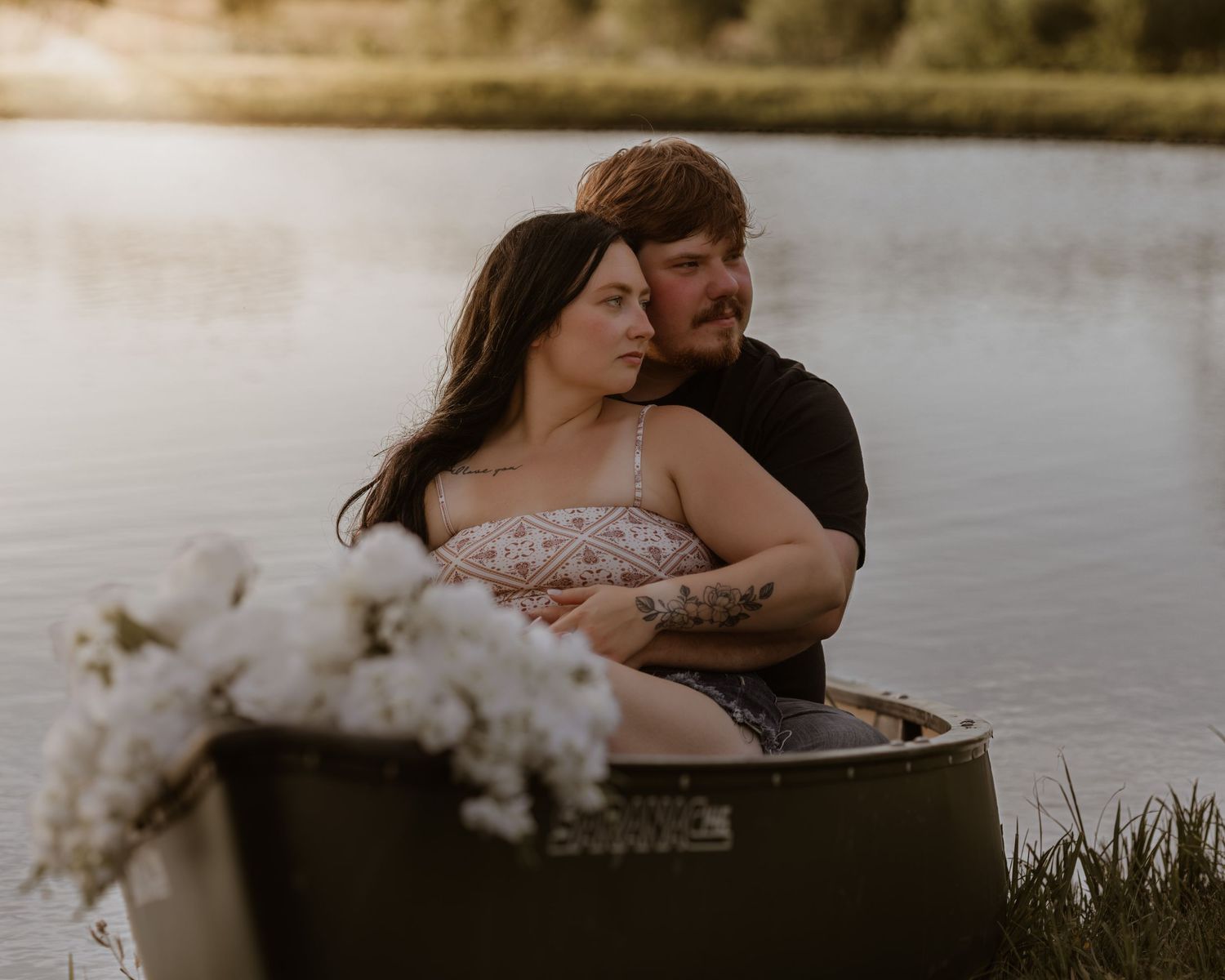 Couple sitting together in a vintage bathtub by a lake at golden hour, surrounded by white flowers.
