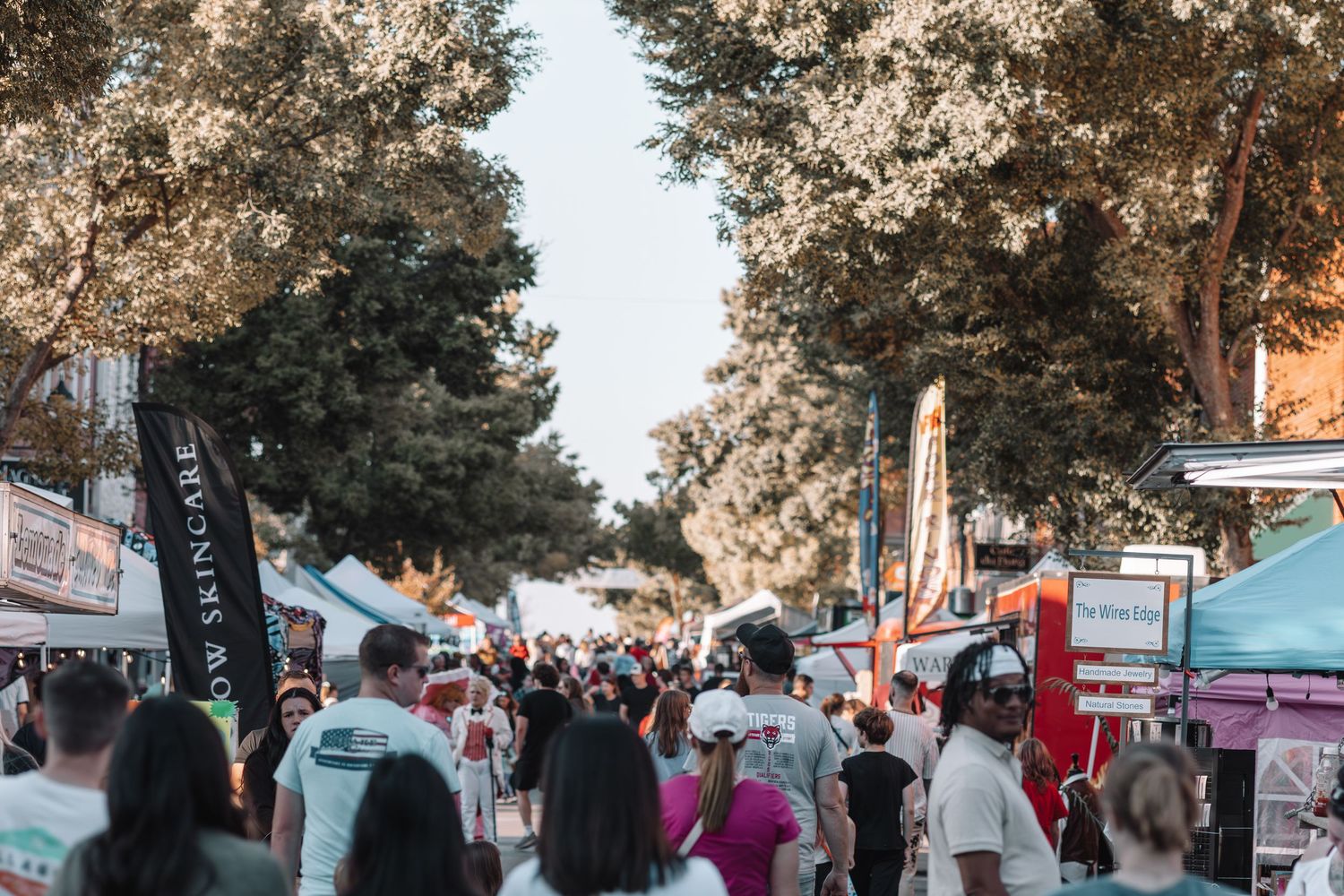 A crowd gathers at an outdoor event with food trucks and vendor tents during sunset under tall pine trees.