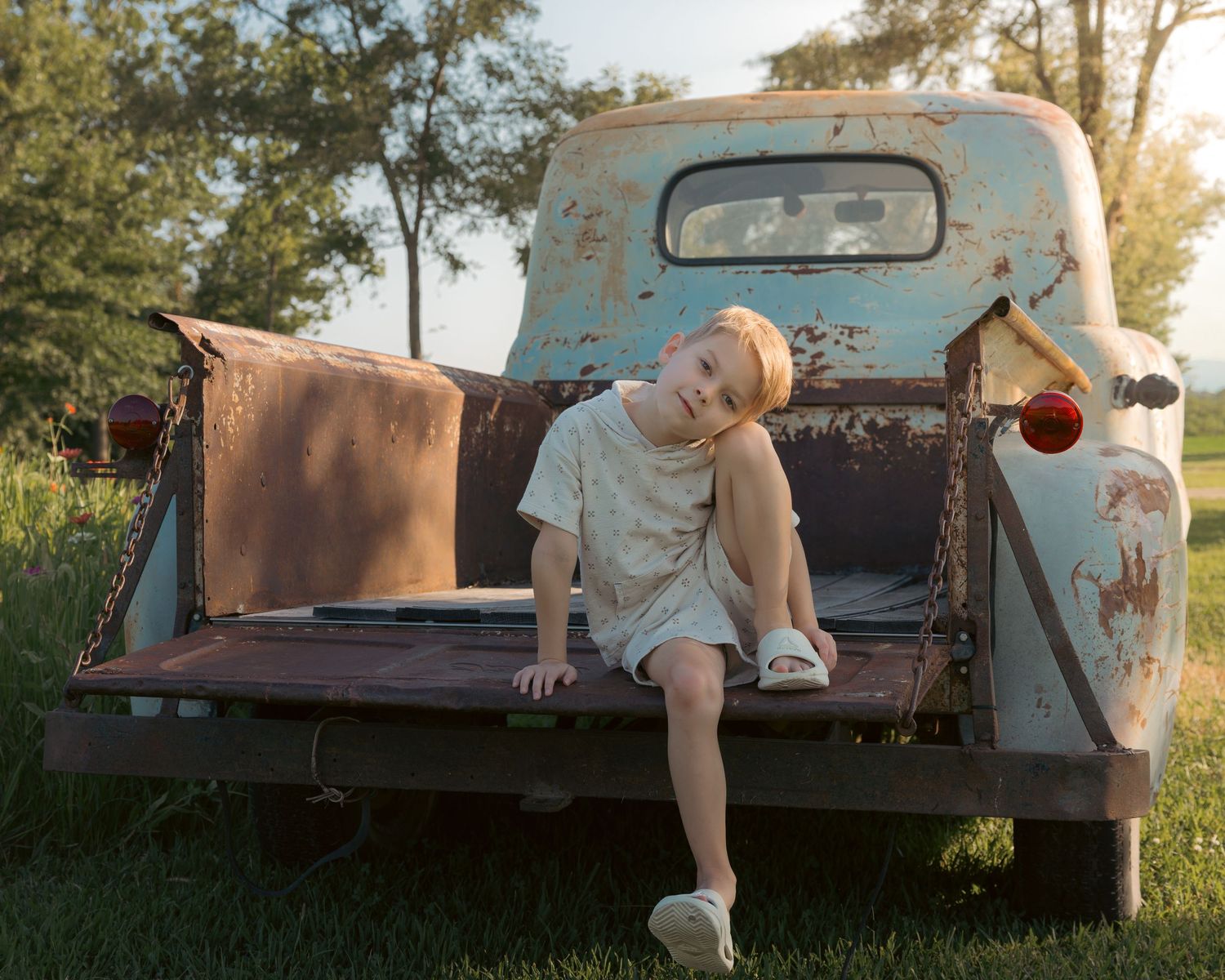 A young child sits on the rusty tailgate of a vintage light blue pickup truck in a sunny green field.