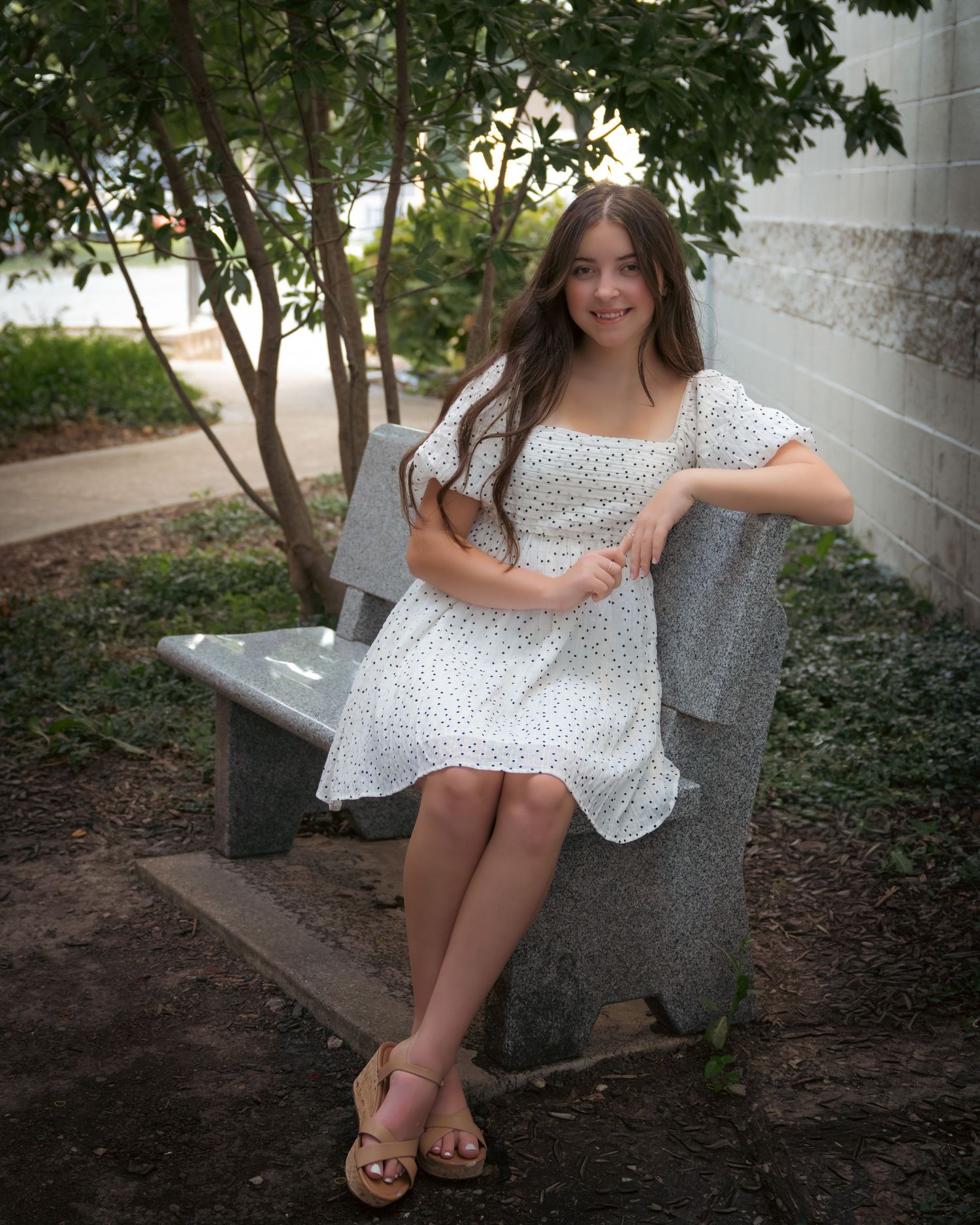 Young woman in white floral dress sitting on stone bench outdoors, smiling, wearing sandals near green trees.