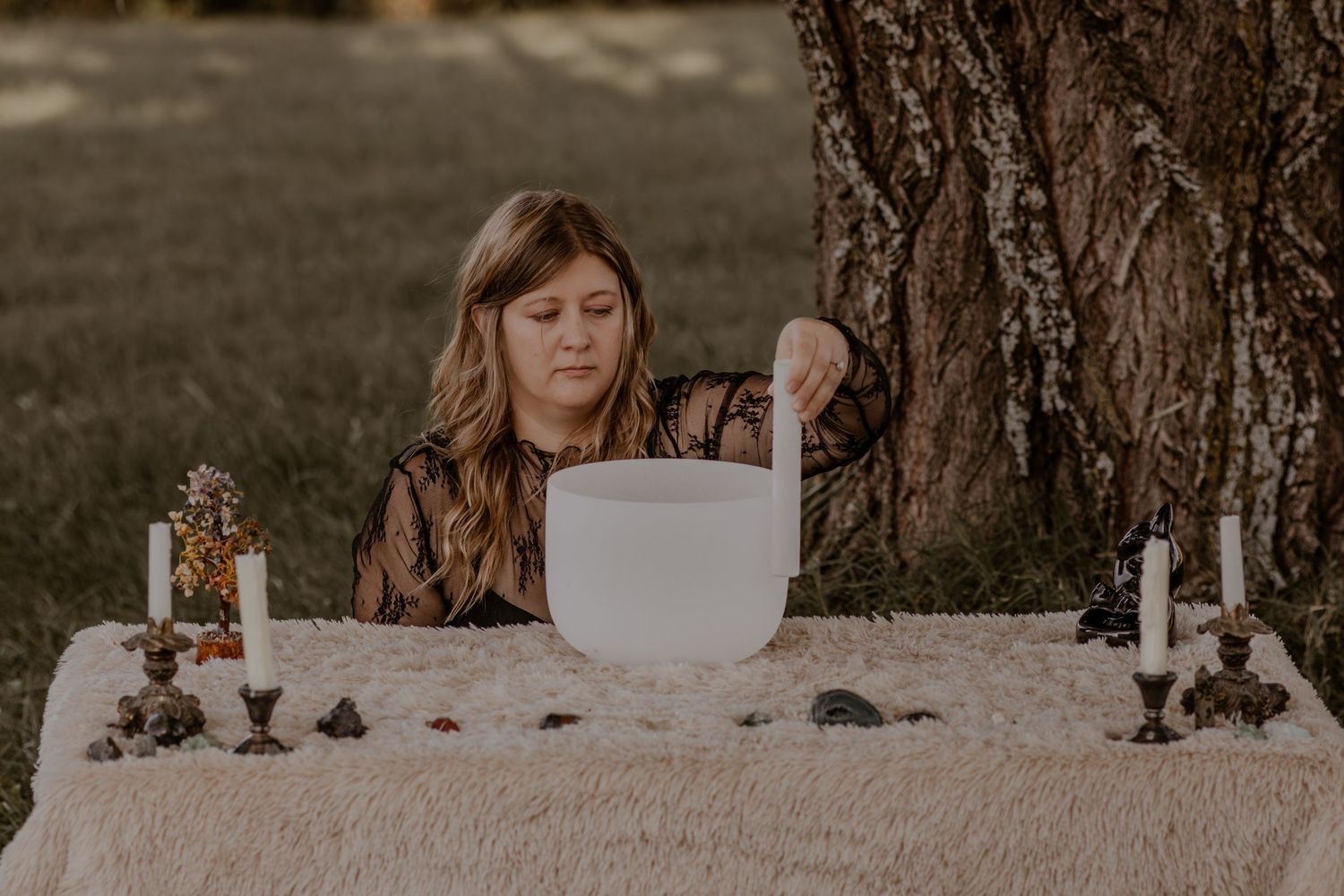 A rustic outdoor table setting with candles and crystals features someone stirring a white ceramic bowl at sunset.