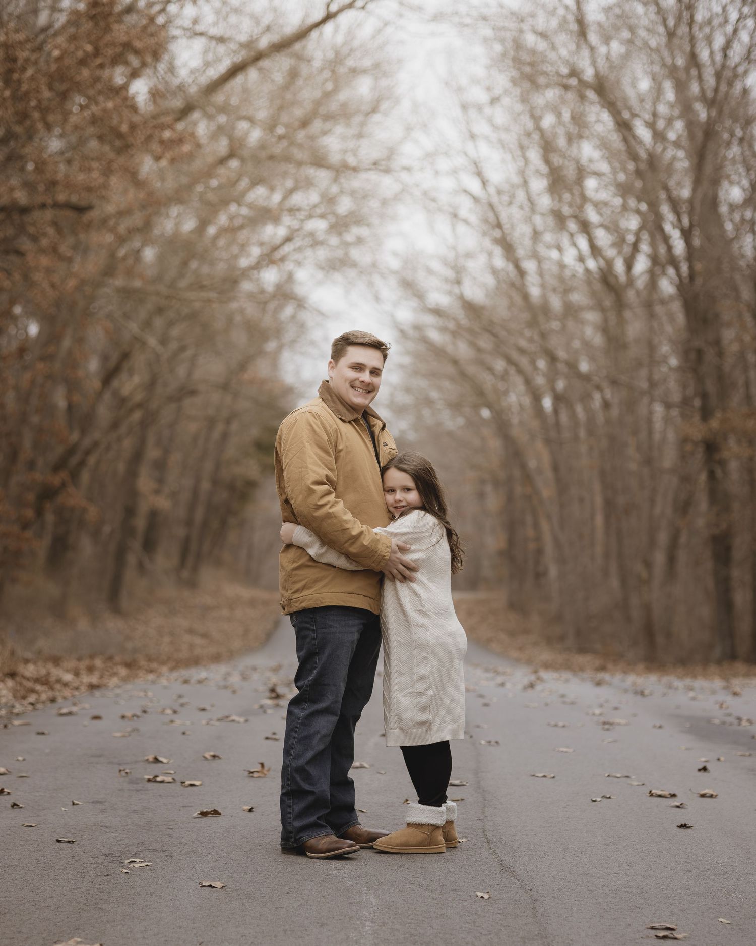 A couple shares a tender moment on a path surrounded by bare autumn trees in a sepia-toned photograph.