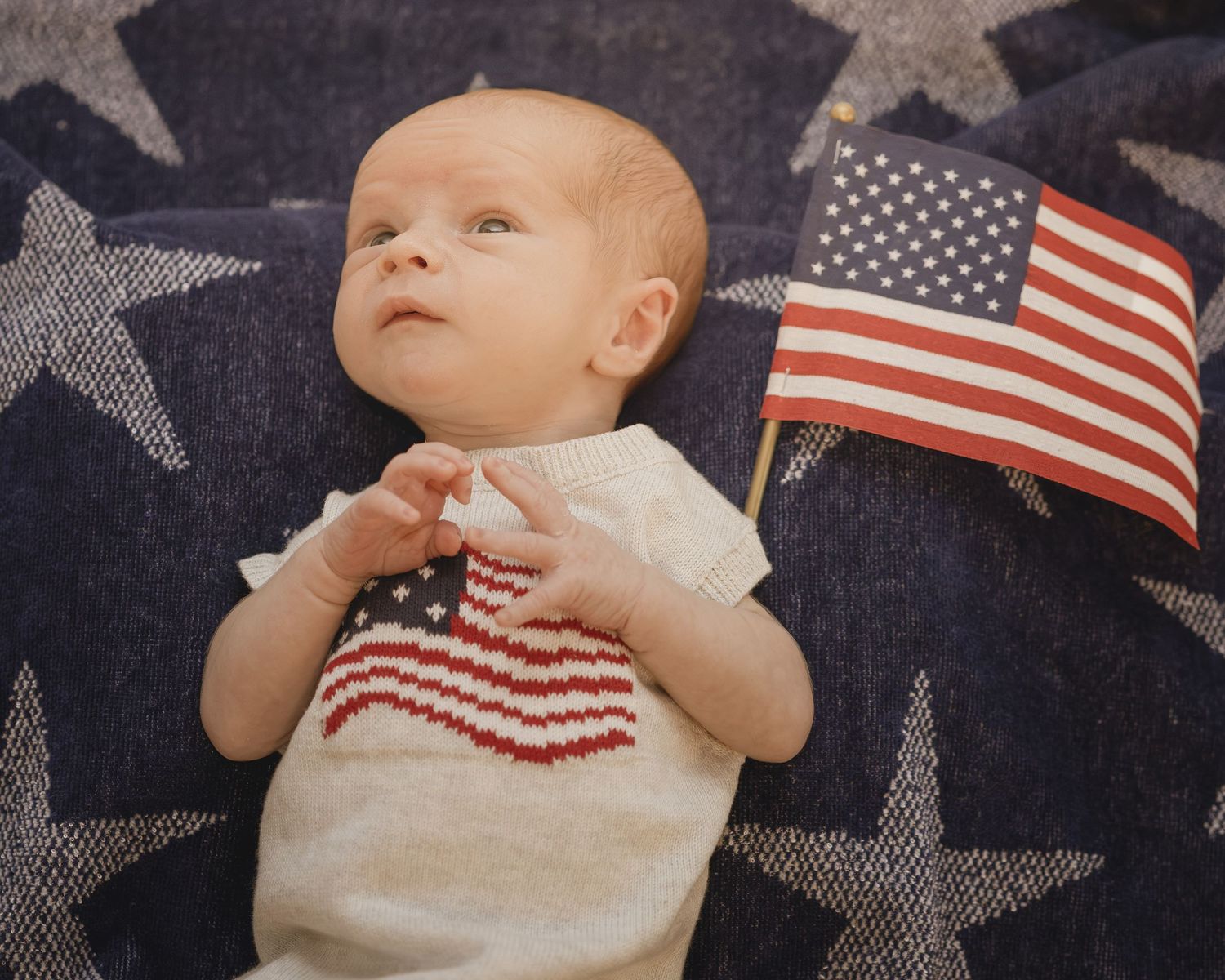 Newborn baby in an American flag onesie lying on a star-patterned blanket with a small US flag.