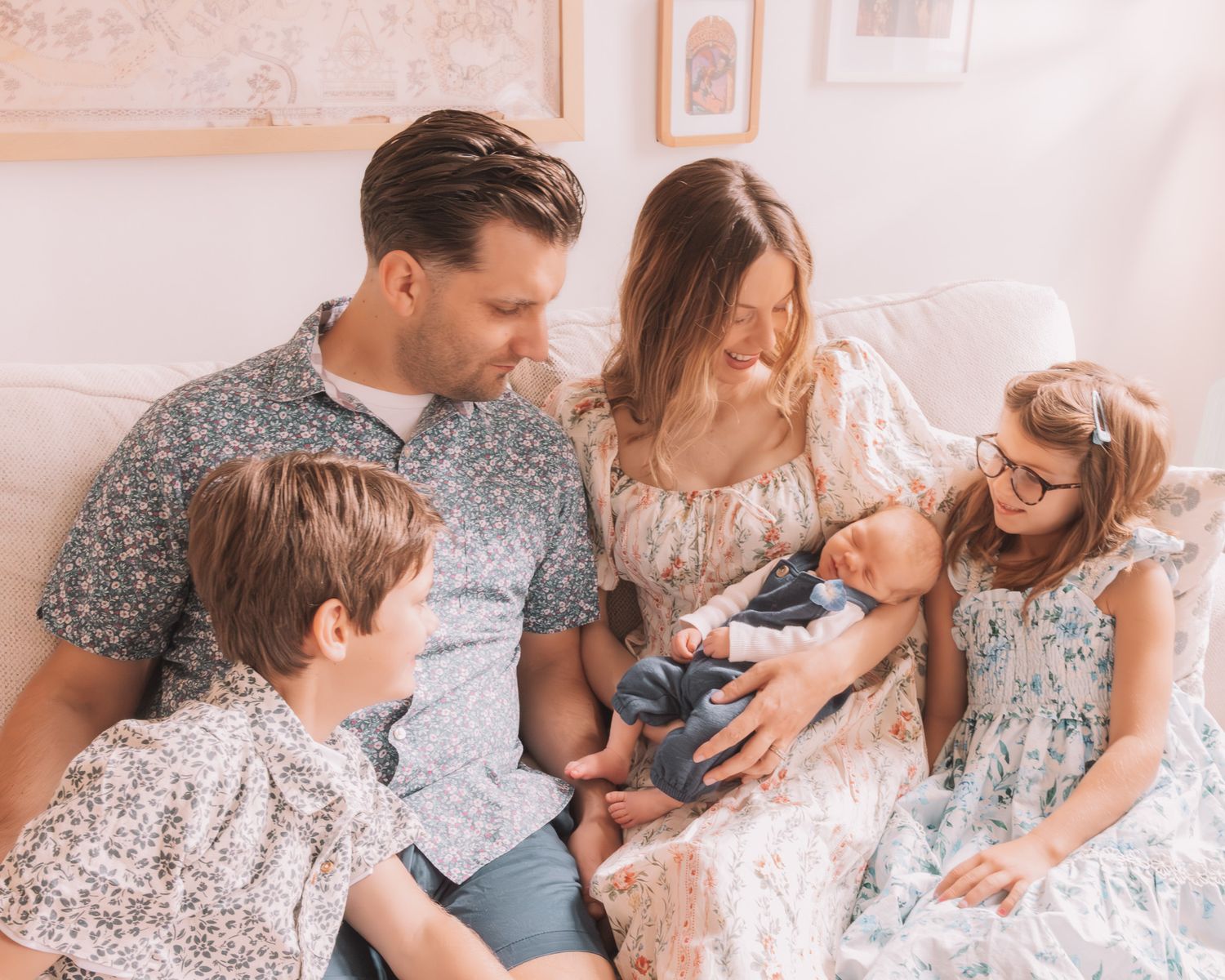 Family of five sitting on a couch admiring their newborn baby in a bright, cozy living room setting.