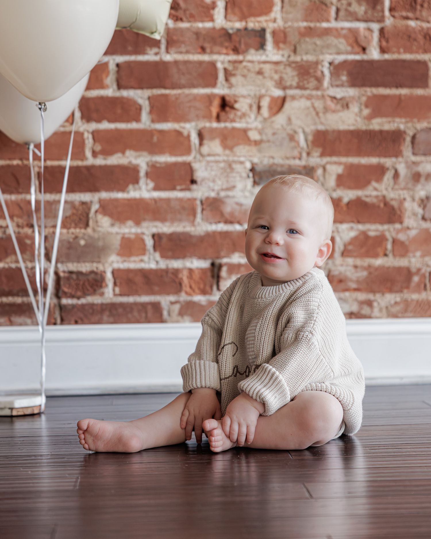 Smiling baby in beige knit romper sits on wood floor near white balloons against rustic brick wall.