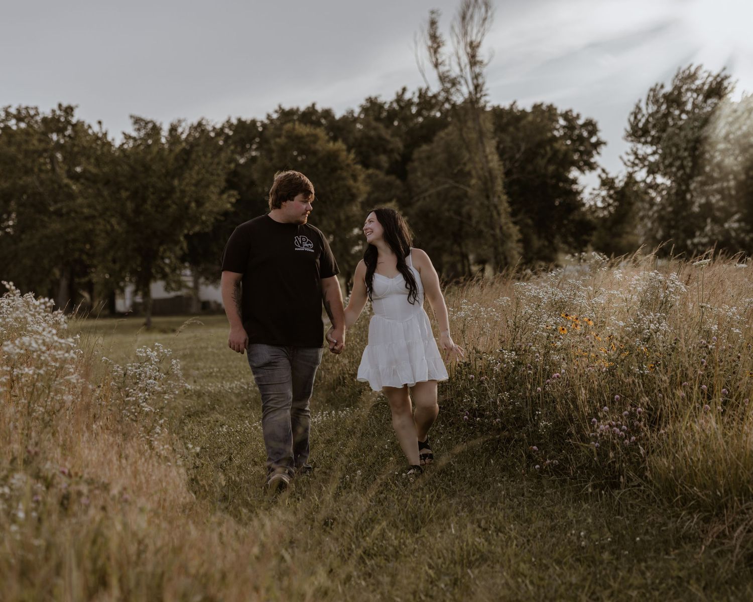 A couple holds hands walking through a wildflower meadow, the woman in a white dress and man in black shirt.