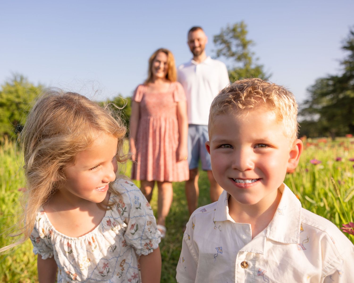 Family portrait with two young children in foreground and parents standing in a sunny green meadow.