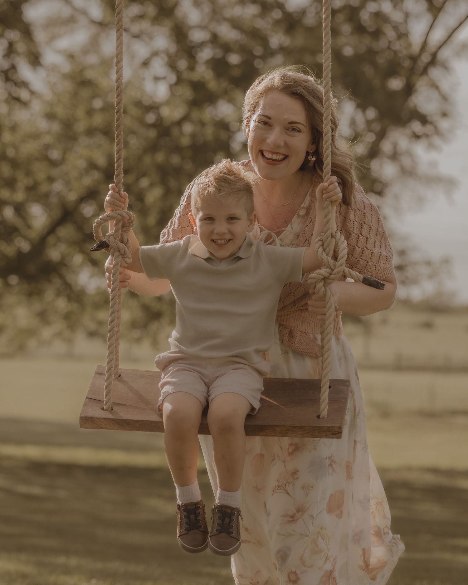 A smiling woman in a floral dress pushes a happy young boy on a wooden rope swing outdoors.