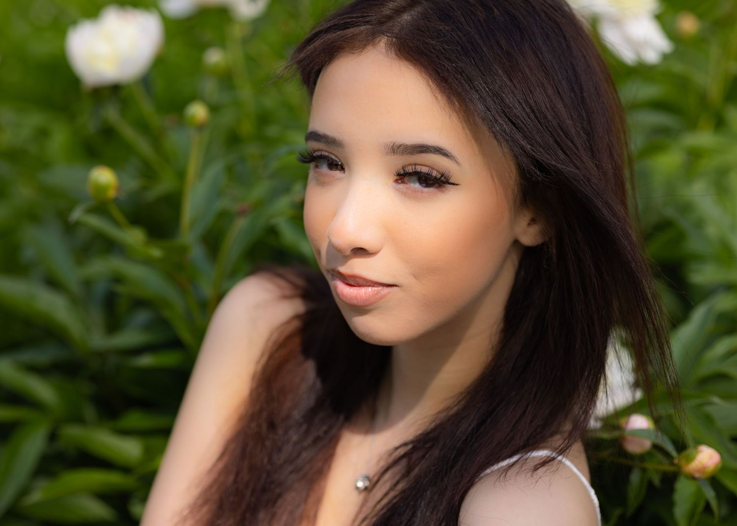 Young woman with dark hair poses outdoors near white peony flowers, wearing a delicate necklace in soft natural light.