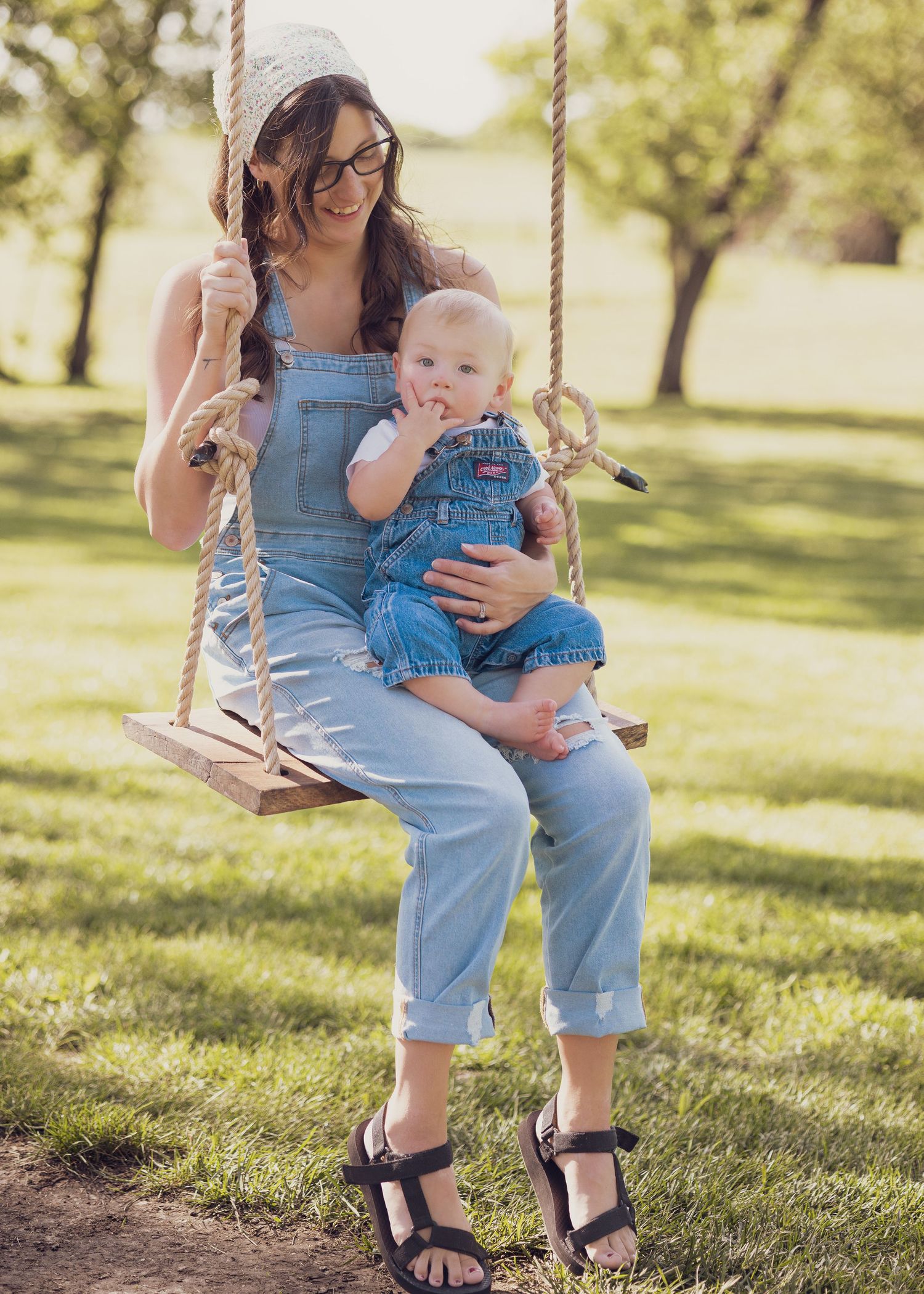 A smiling woman in denim overalls sits on a rope swing holding a baby in matching overalls outdoors.