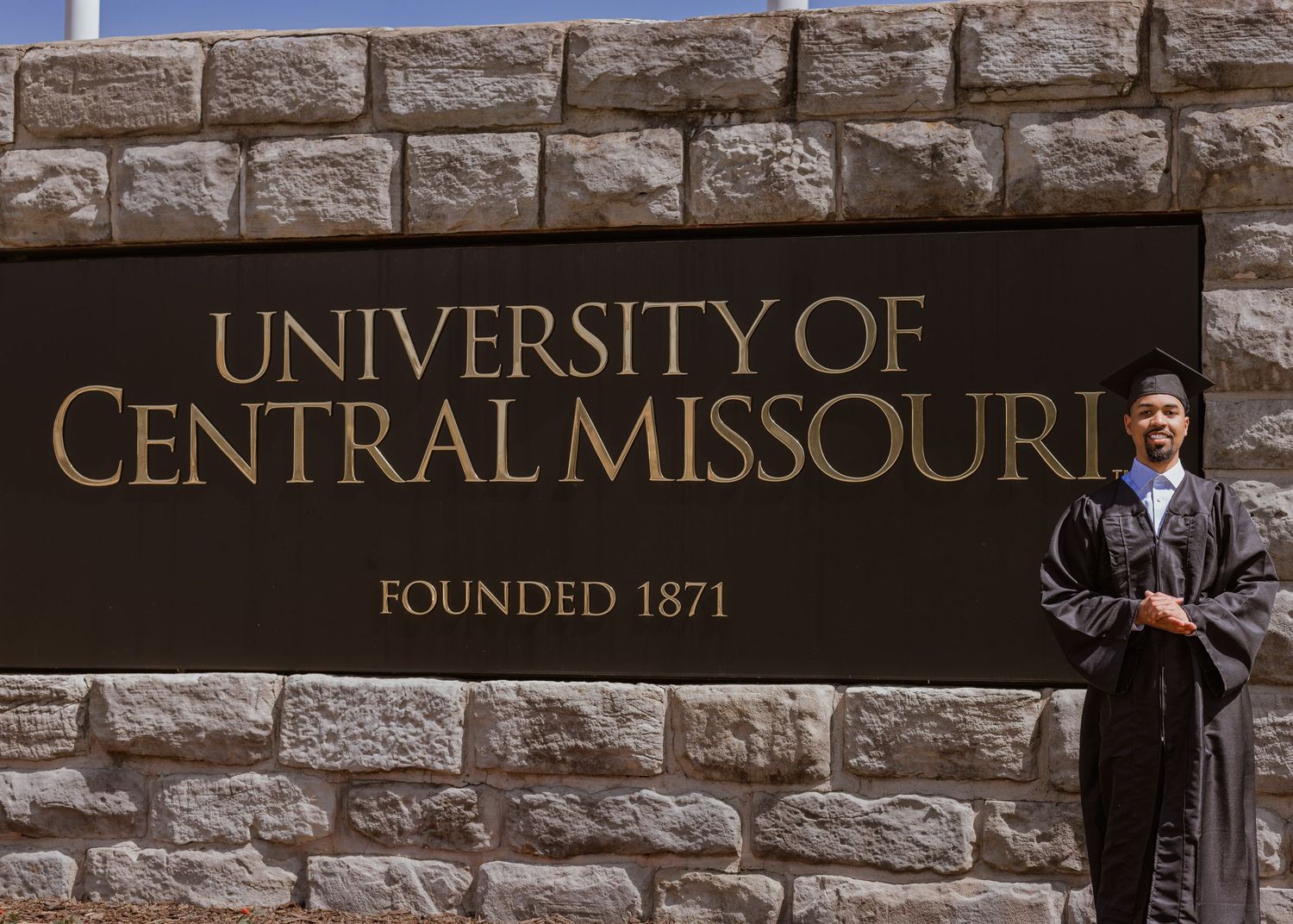 Graduate in cap and gown posing by University of Central Missouri stone sign, founded 1871.