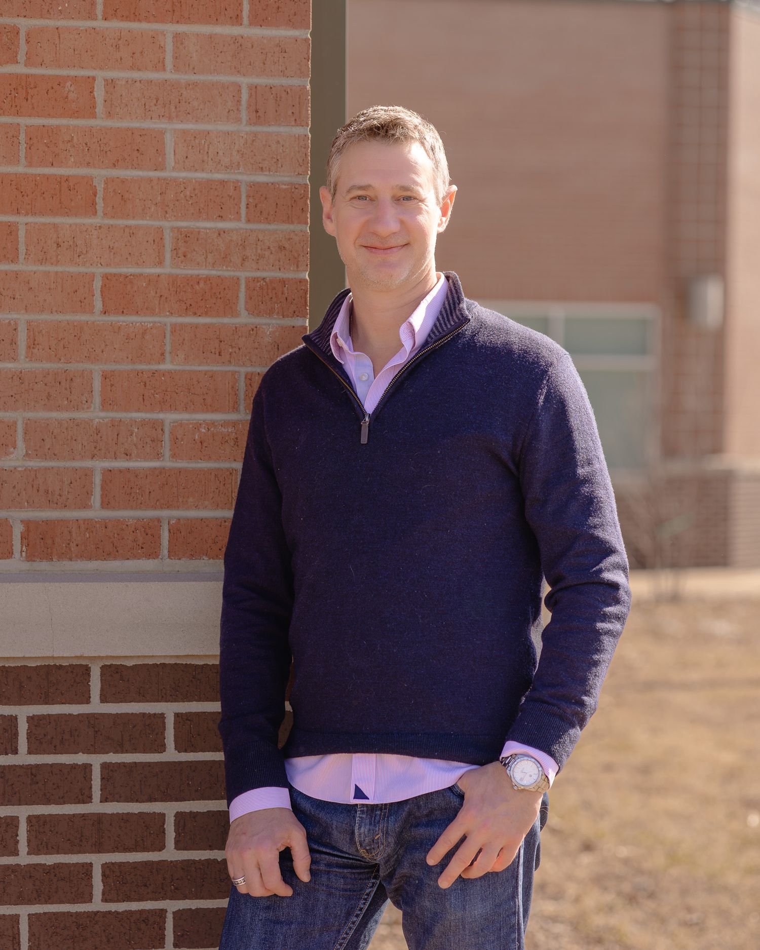 Man in navy quarter-zip sweater leaning against brick wall outdoors, smiling confidently in casual professional attire.