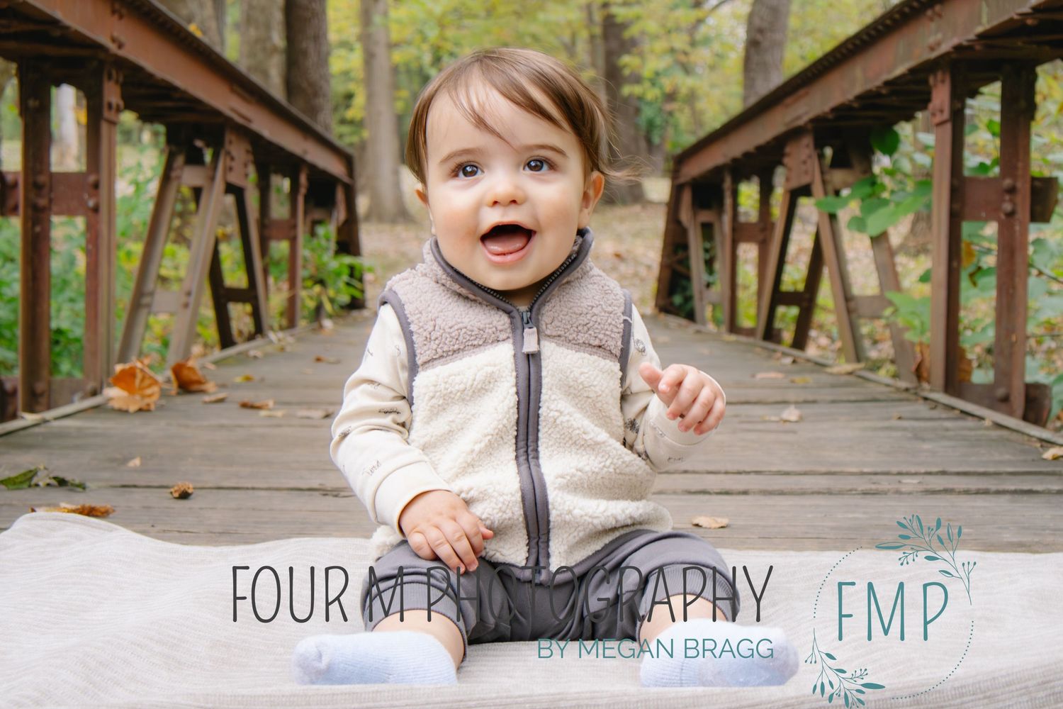 A toddler wearing a gray zip-up sweater sits excitedly on a wooden bridge during autumn.