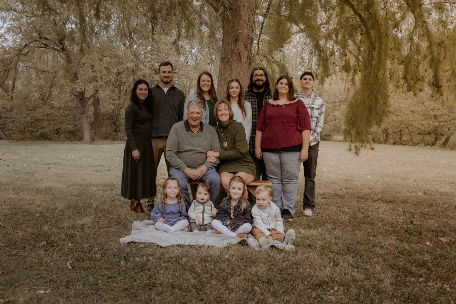 A large multigenerational family poses together outdoors on a blanket beneath tall trees during a fall portrait session.