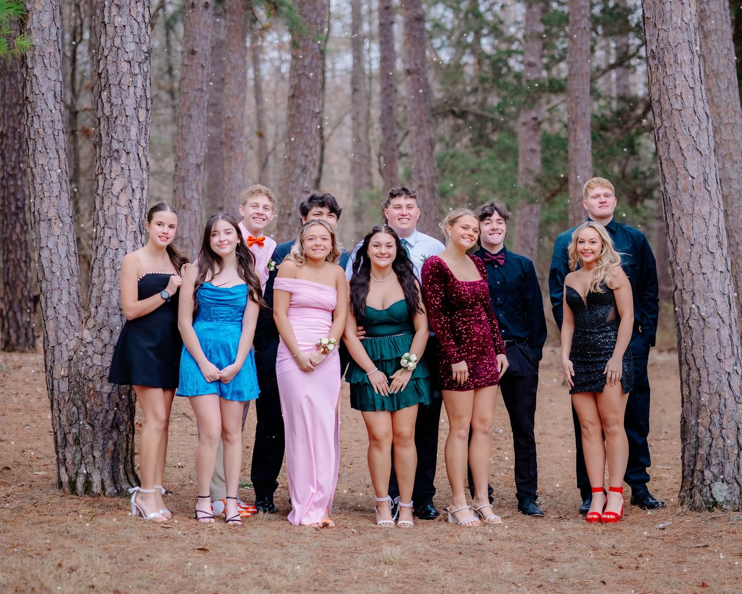 A group of people standing in a row against tall pine trees wearing formal attire and casual clothing.