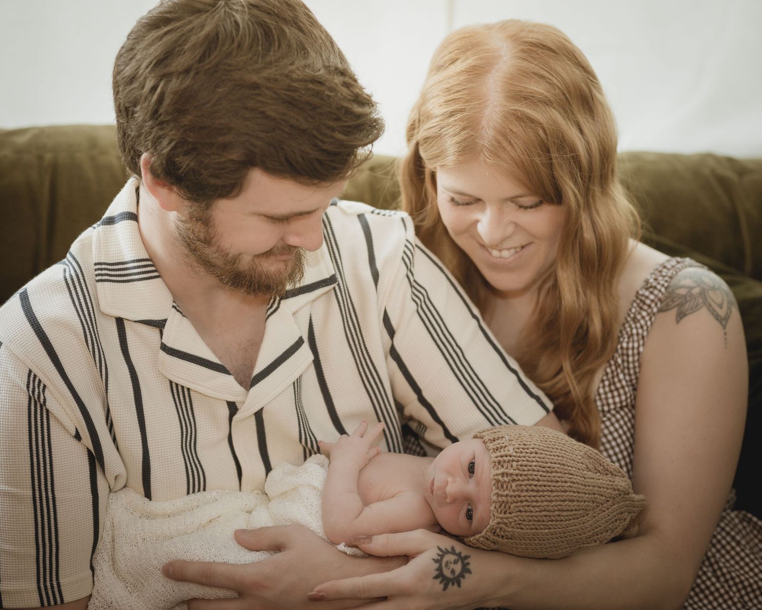 Young parents smiling lovingly at their newborn baby held in dads arms on a couch.