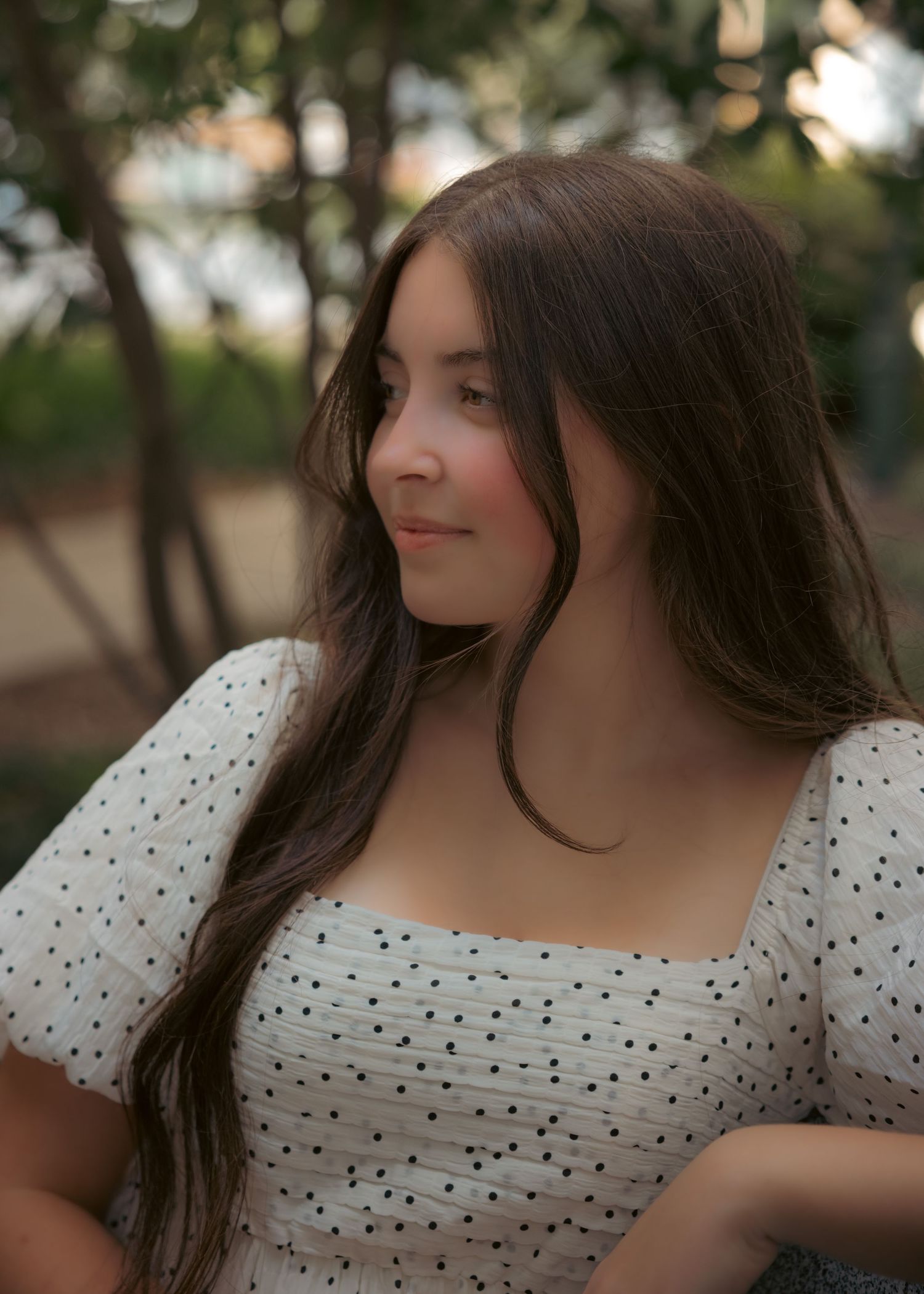 Young woman with long brown hair wearing a white polka dot dress, smiling outdoors among trees at sunset.