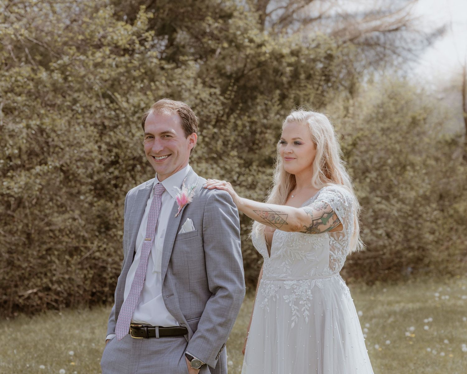 Bride in white lace gown places hand on groom's shoulder in gray suit during outdoor wedding portrait.