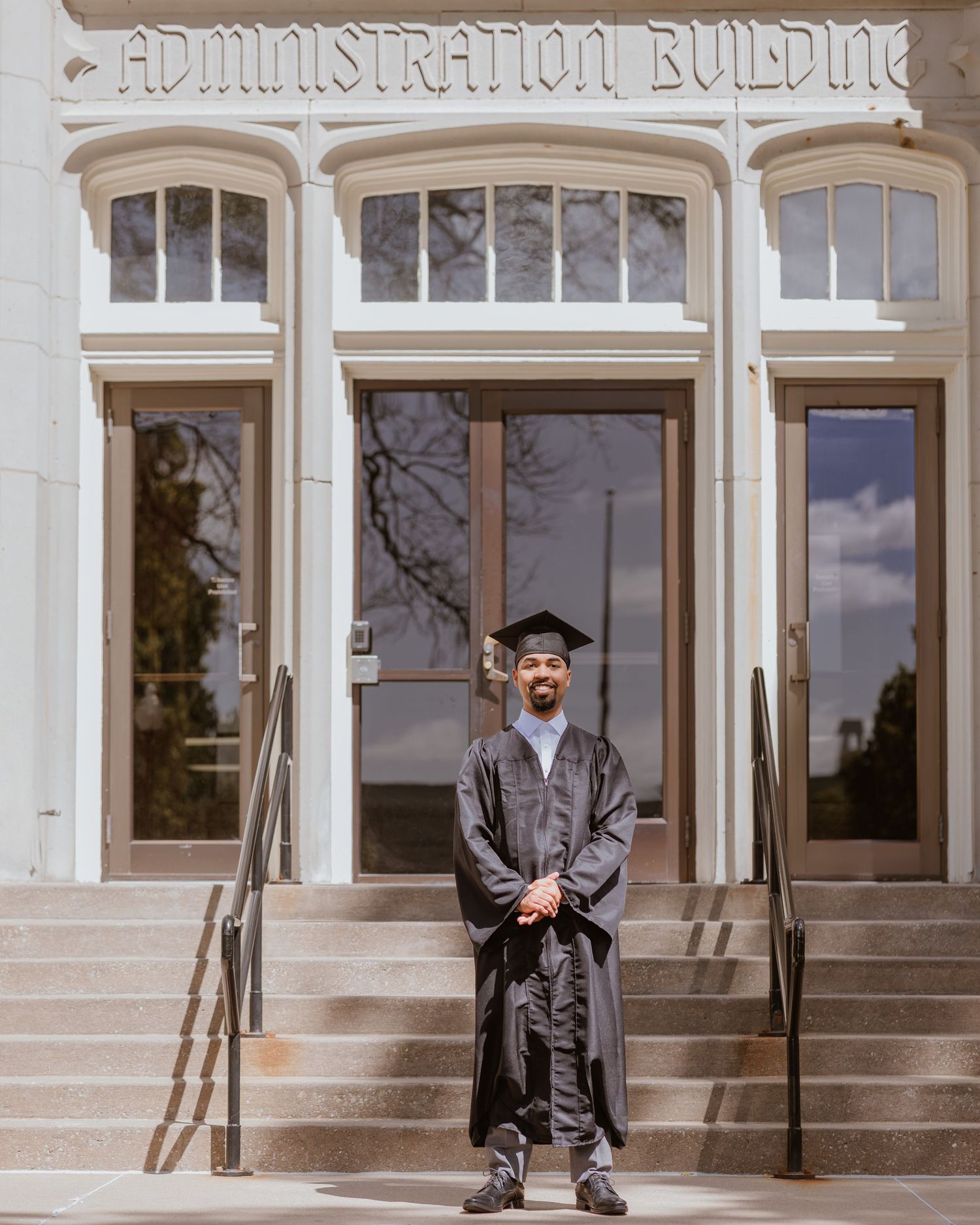 Graduate in cap and gown stands proudly on steps of Administration Building on college campus graduation day.