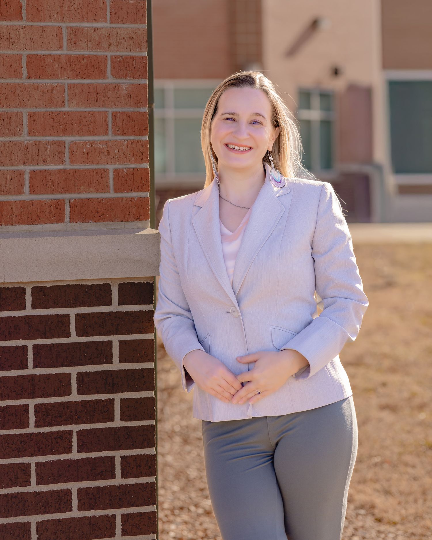 Smiling blonde woman in light gray blazer and pants leaning against brick wall outdoors in professional photo.