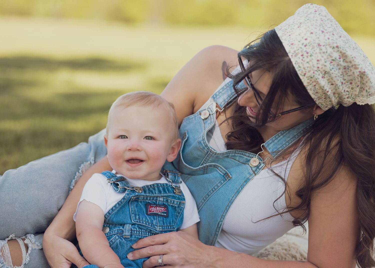 Laughing baby boy in denim overalls with mother lying in grass on sunny day.