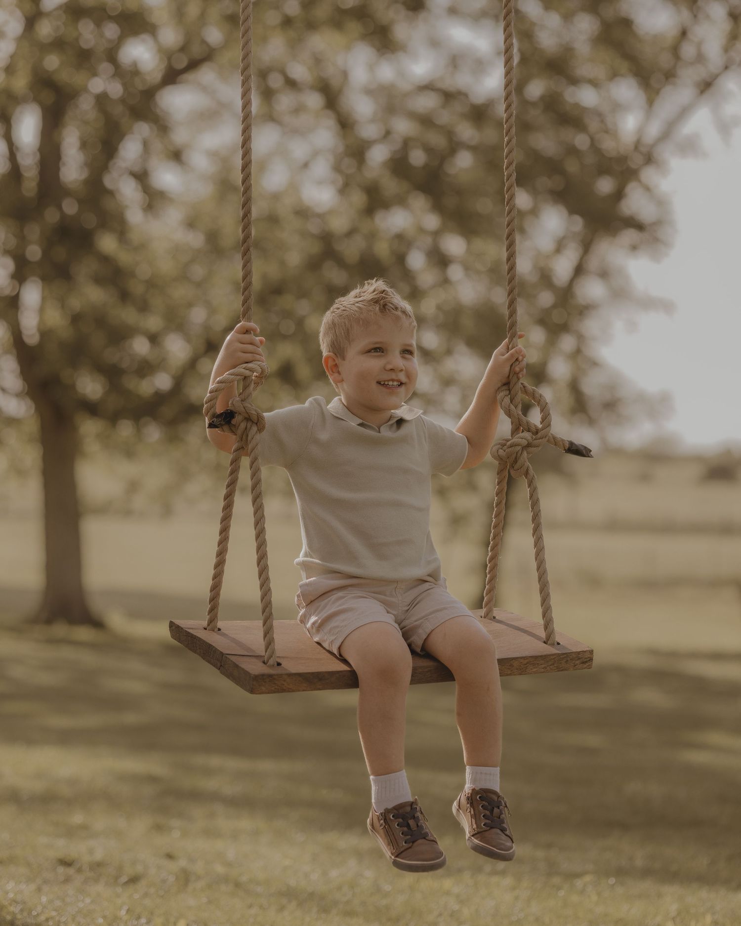 A cheerful young boy in a light shirt and shorts swings on a wooden rope swing in a grassy field.