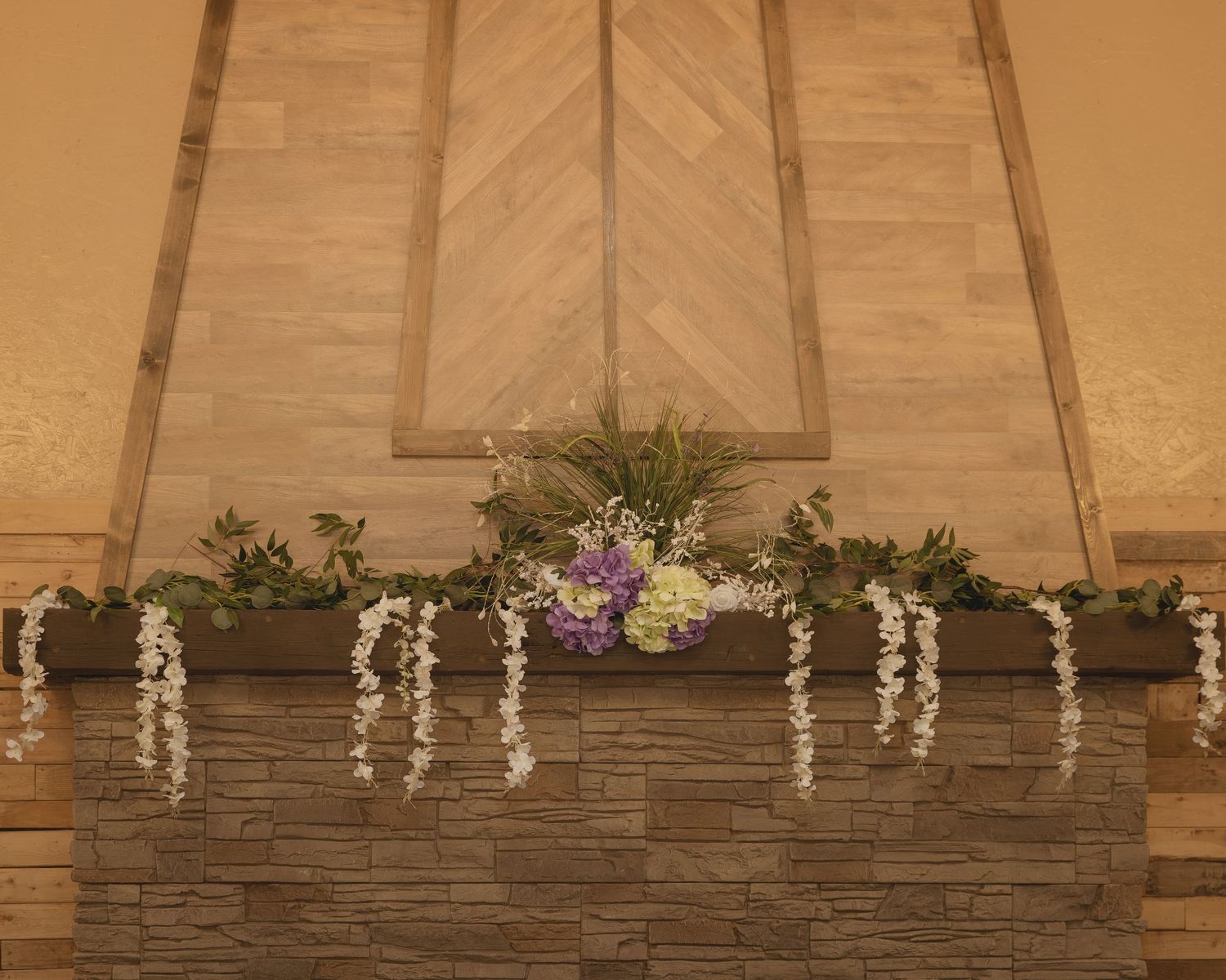 Floral garland with white wisteria and purple blooms decorates a stone fireplace mantel beneath a wooden chevron wall.
