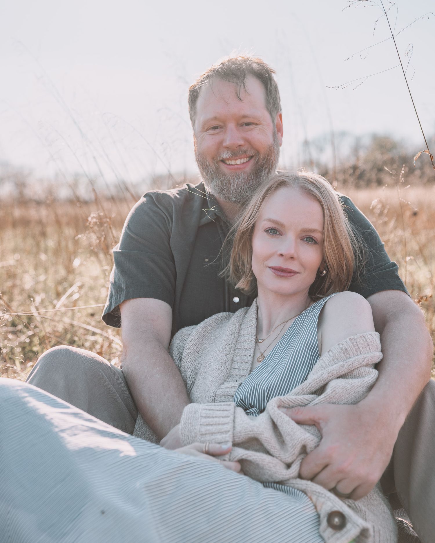 A smiling couple sitting together in a golden field, the man with a beard embracing a fair-skinned woman in a cozy cardigan.