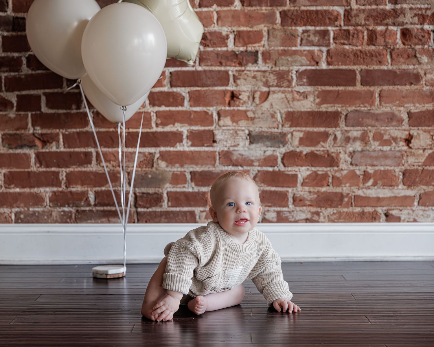 Baby in cream knit romper crawling on hardwood floor near white balloons against rustic brick wall.