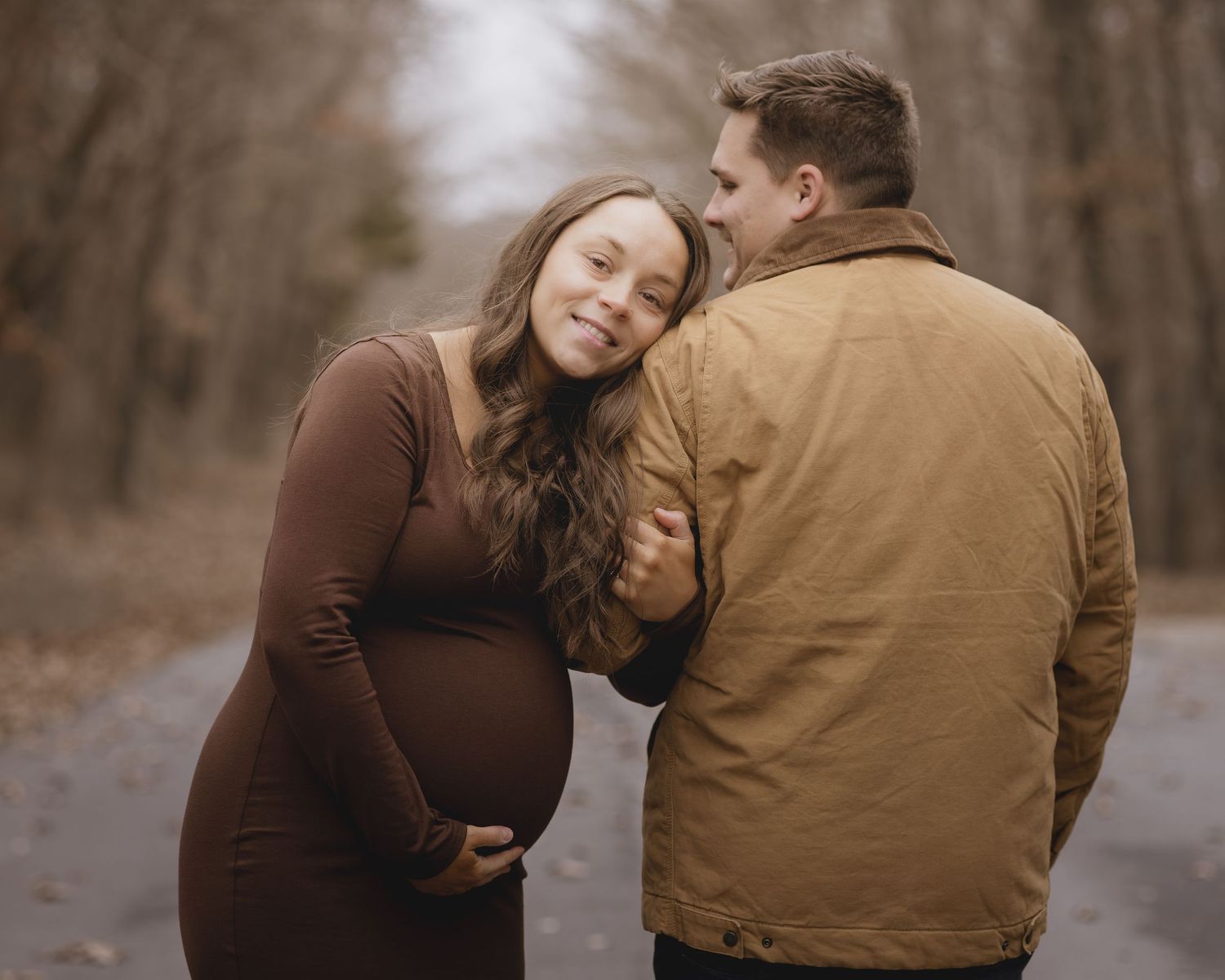 Pregnant couple sharing a tender moment on a forest path in autumn, woman in brown dress cradling baby bump.